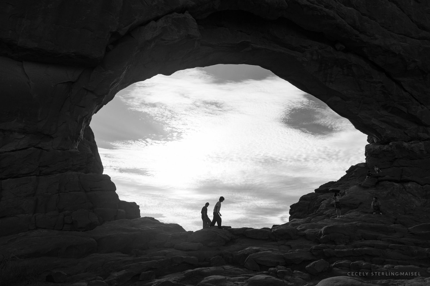 Black and white: two figures stand silhouetted in the mouth of a great sandstone arch against a bright clouded sky.