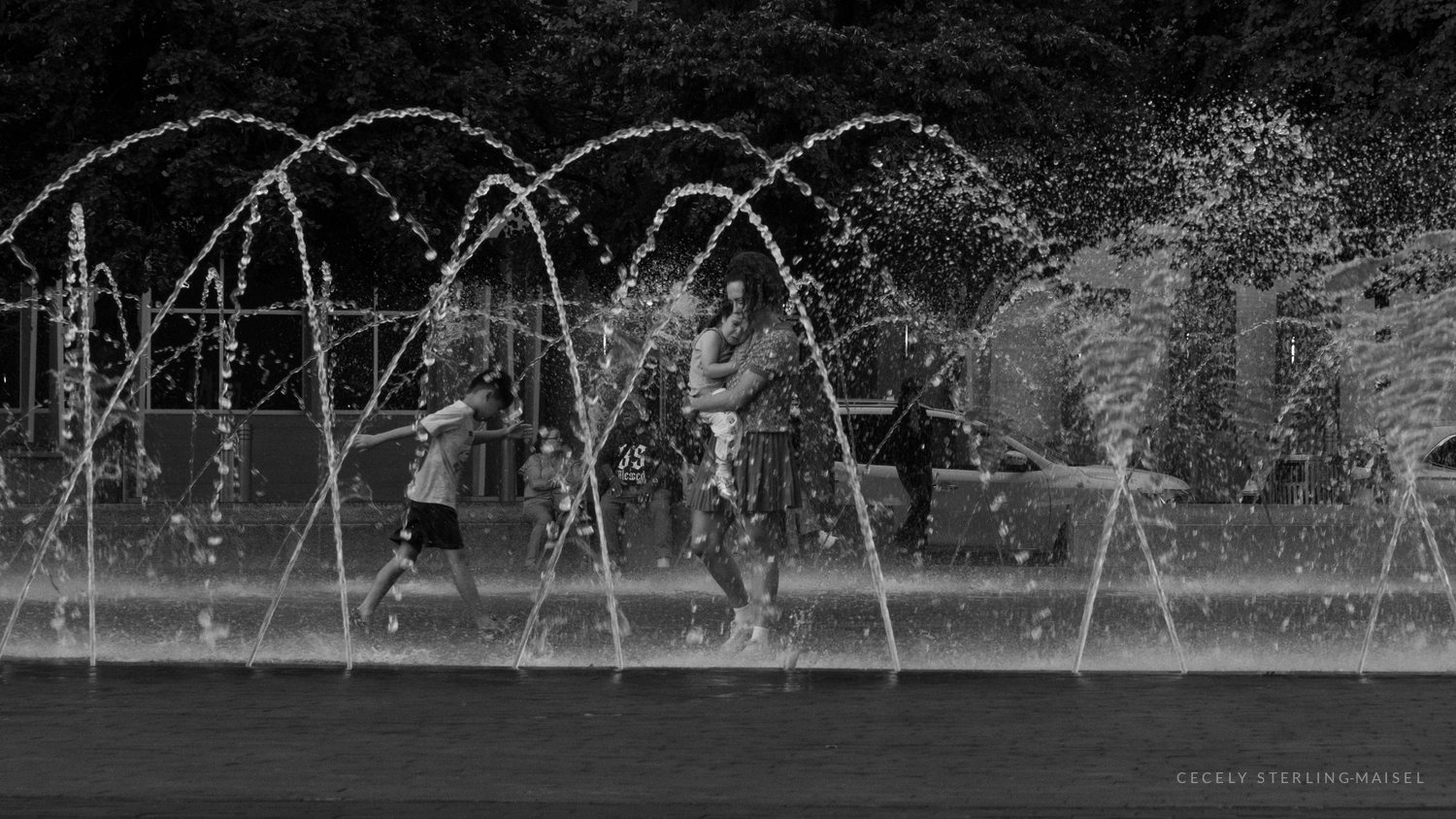 Black and white photo of children and an adult playing among the arcing jets of a public fountain, water spraying in bright streaks.