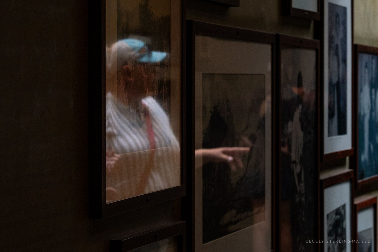 A person in a blue cap and striped shirt, reflected in the glass of a framed print on a gallery wall.