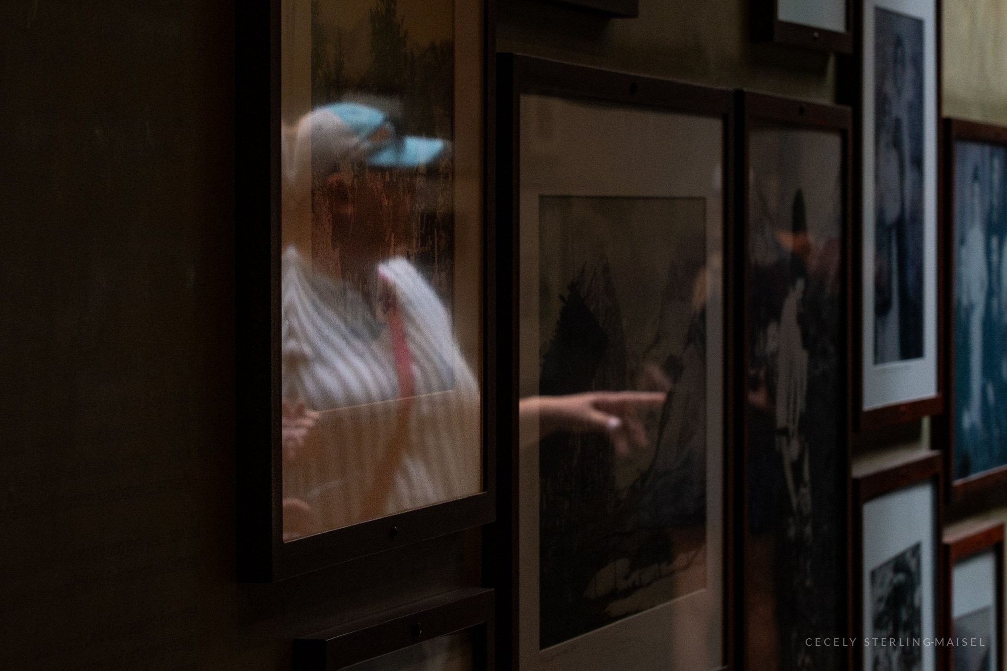 A person in a blue cap and striped shirt, reflected in the glass of a framed print on a gallery wall.