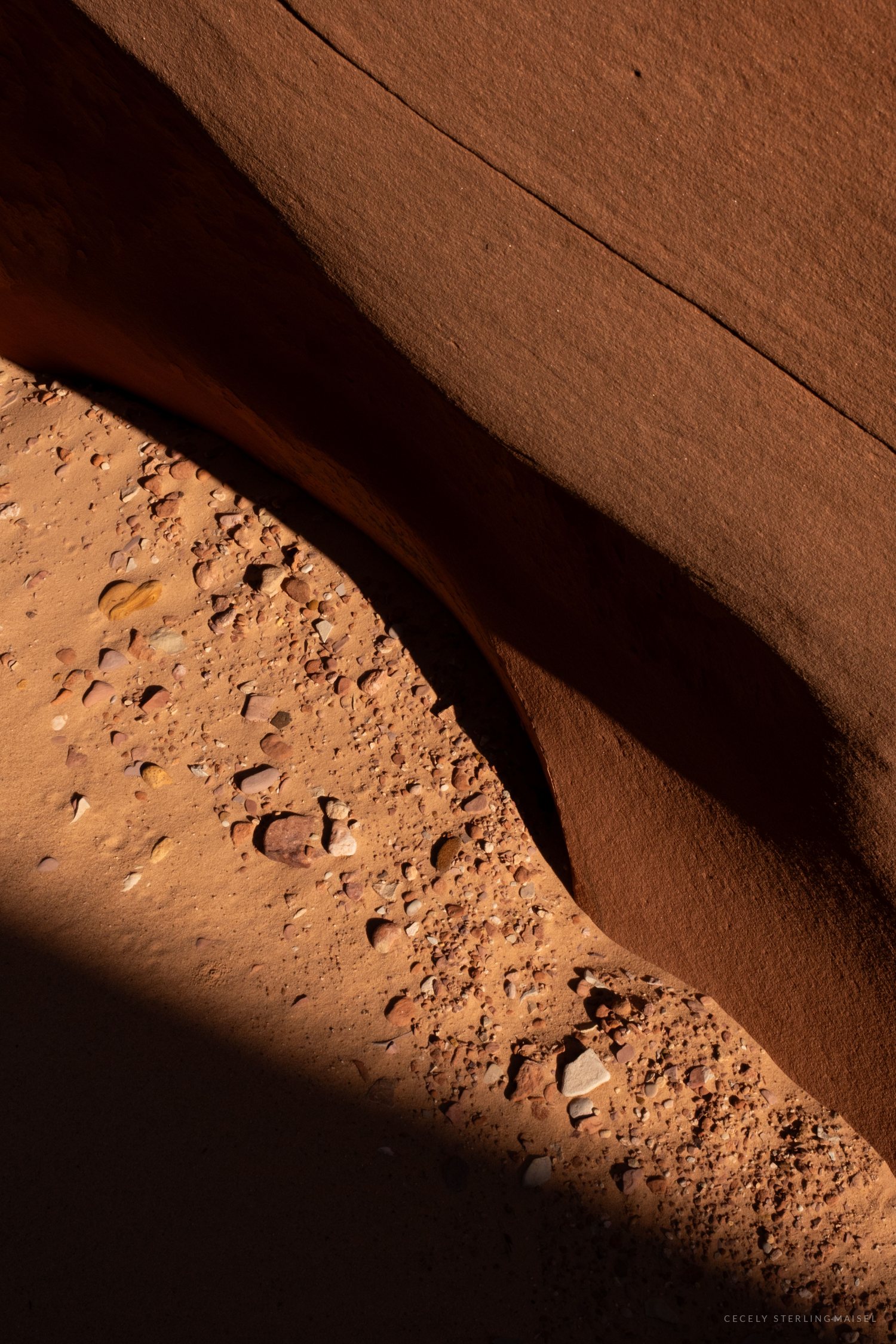 Close-up of sandstone canyon rock, showing a smooth curved overhang casting a dark shadow over a gravel-covered floor
