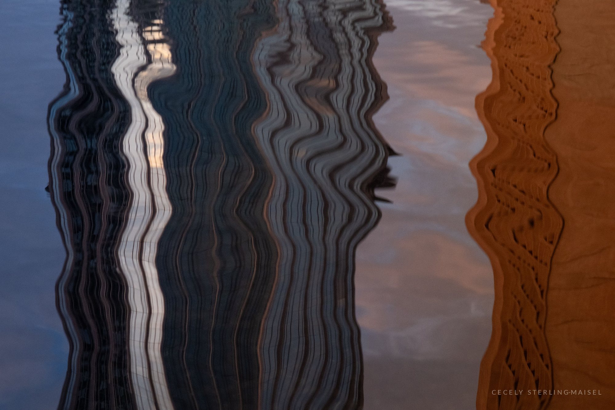 Close-up of water reflecting a glass tower and a warm brick facade, the vertical lines stretched into wavy ribbons.