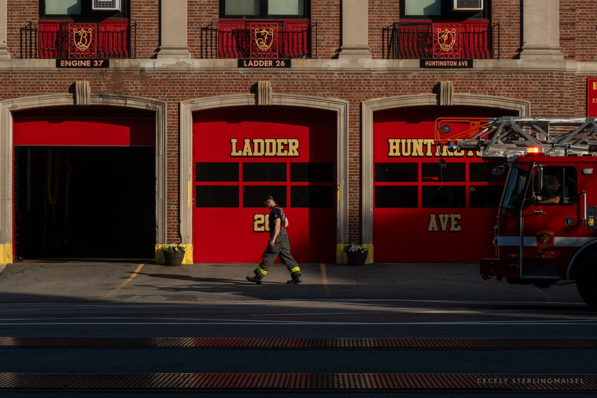 A firefighter walking past the red bay doors of a brick fire station, an engine parked at the right.