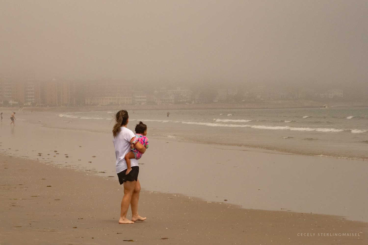 A woman carrying a small child at the water's edge, a hazy shoreline of buildings behind them.