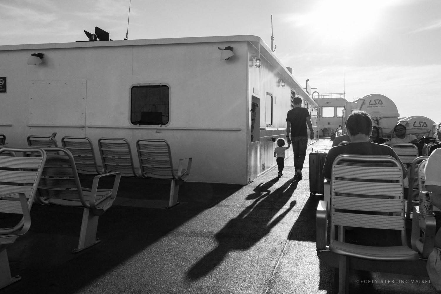 Black and white ferry deck with empty chairs and long shadows thrown by low sun and standing figures.