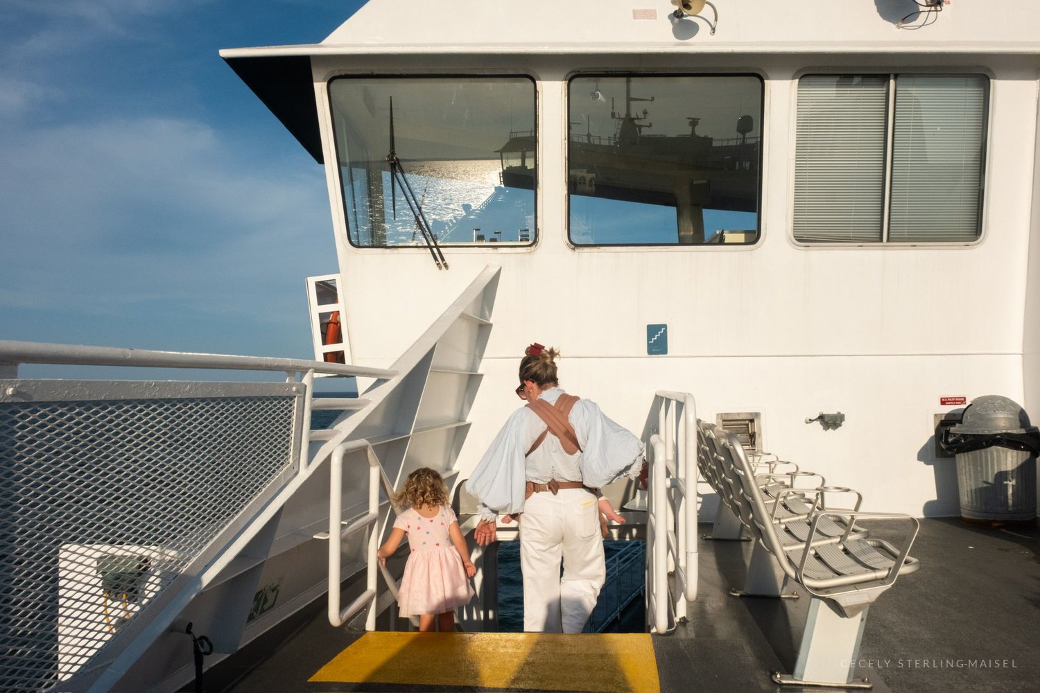 A woman in white leading a small child up the stairs of a ferry against a bright blue sky.