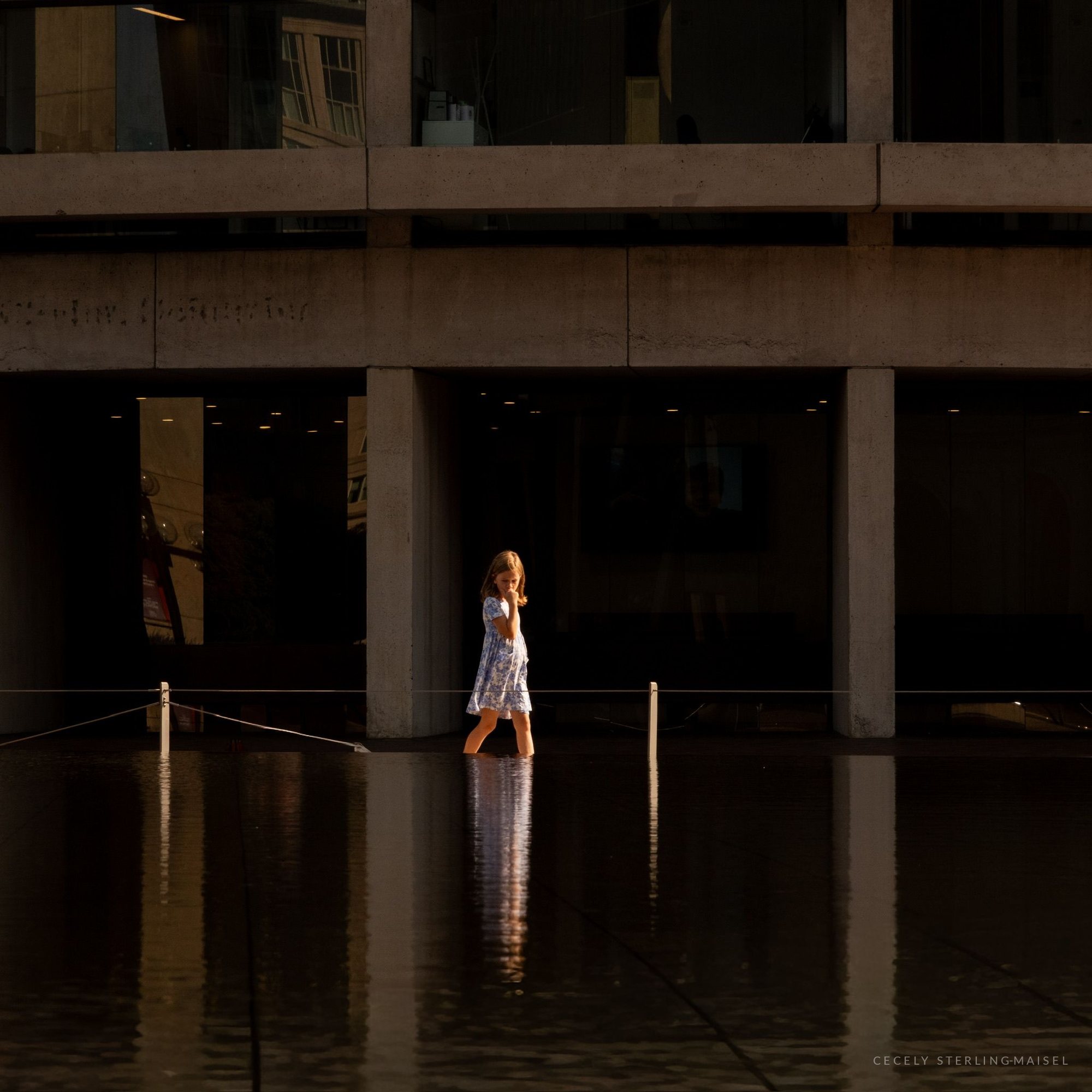 A small figure in white crossing a shallow reflecting pool, framed by the deep shadow of a concrete colonnade.