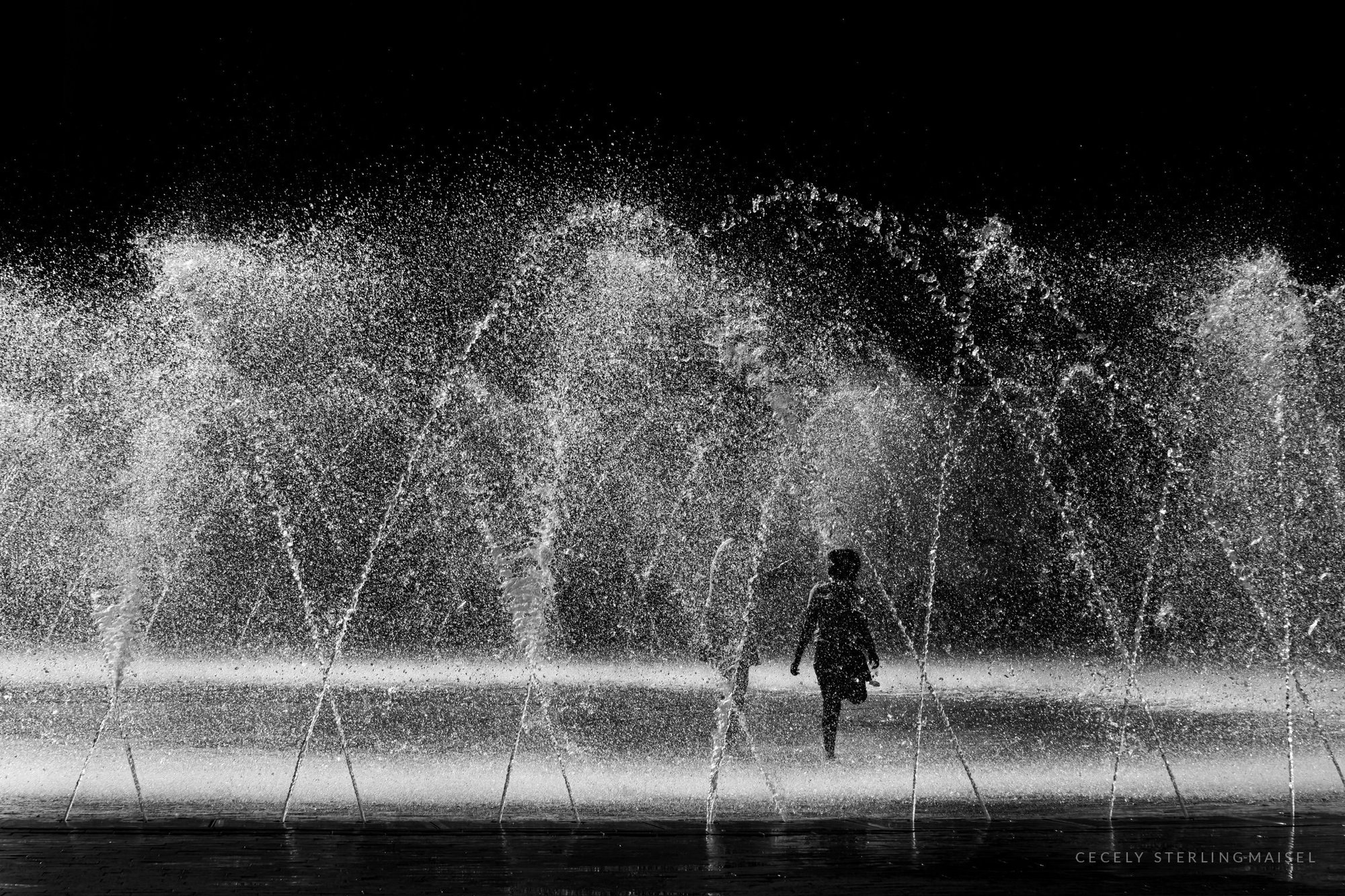 A child running through tall arcs of backlit fountain spray against a dark background.