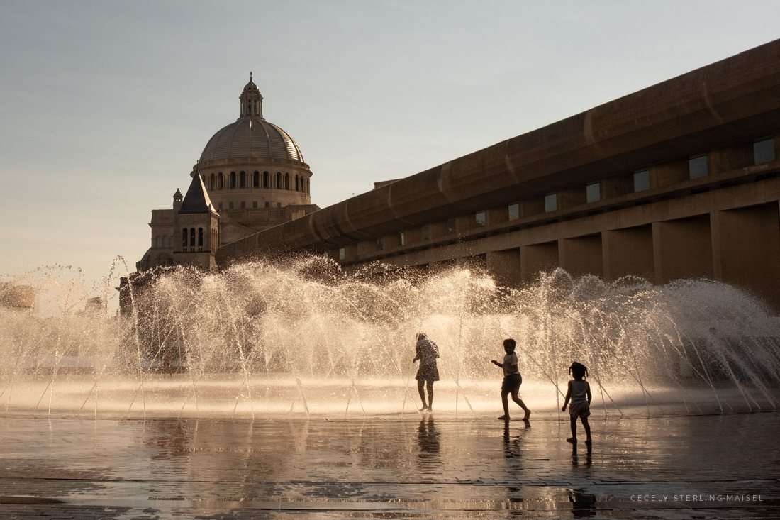 Children running through arcing jets of fountain water, a domed church behind them in warm evening light.