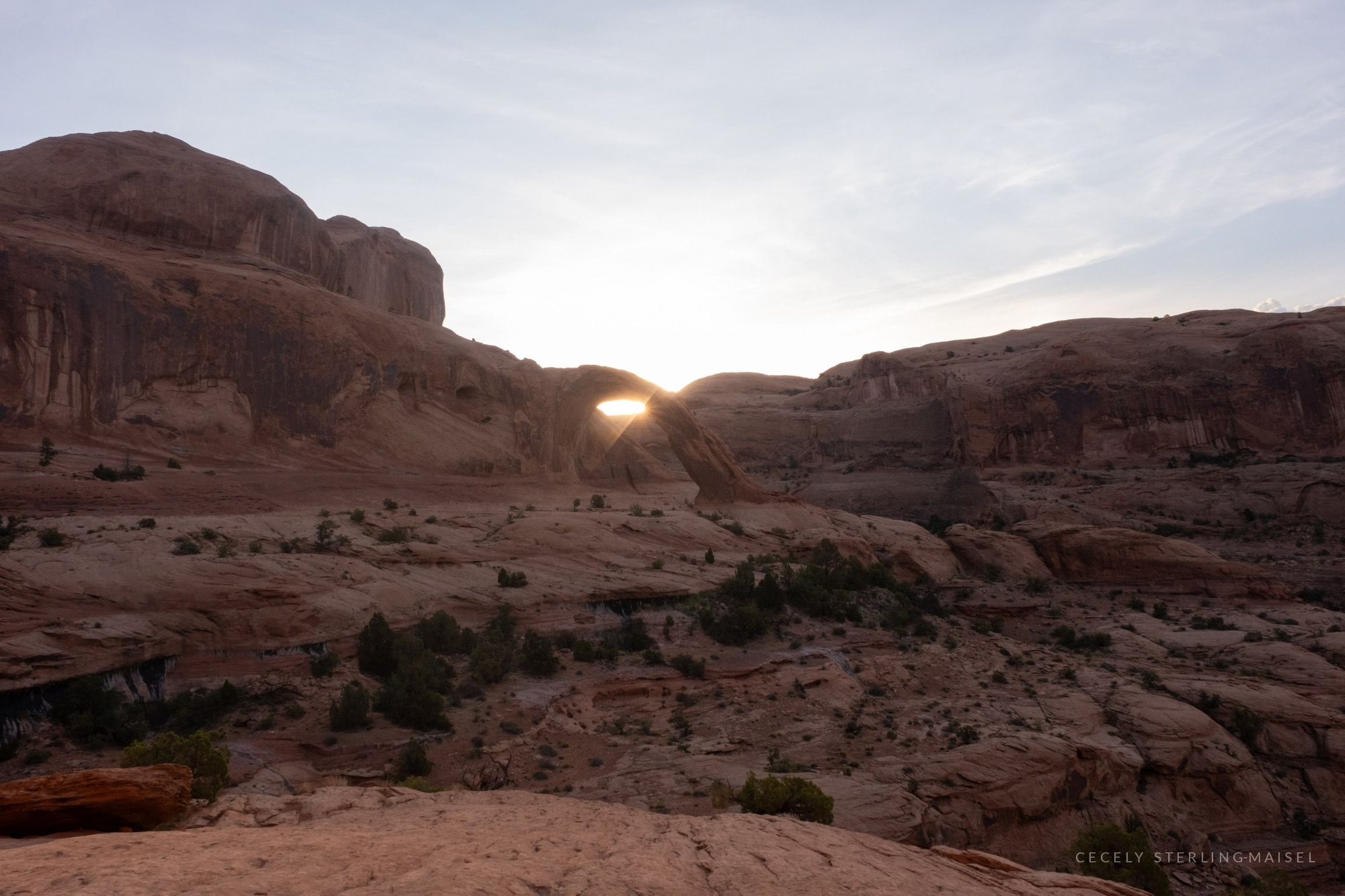 The sun cresting a low sandstone ridge above a wide slickrock basin scattered with juniper.