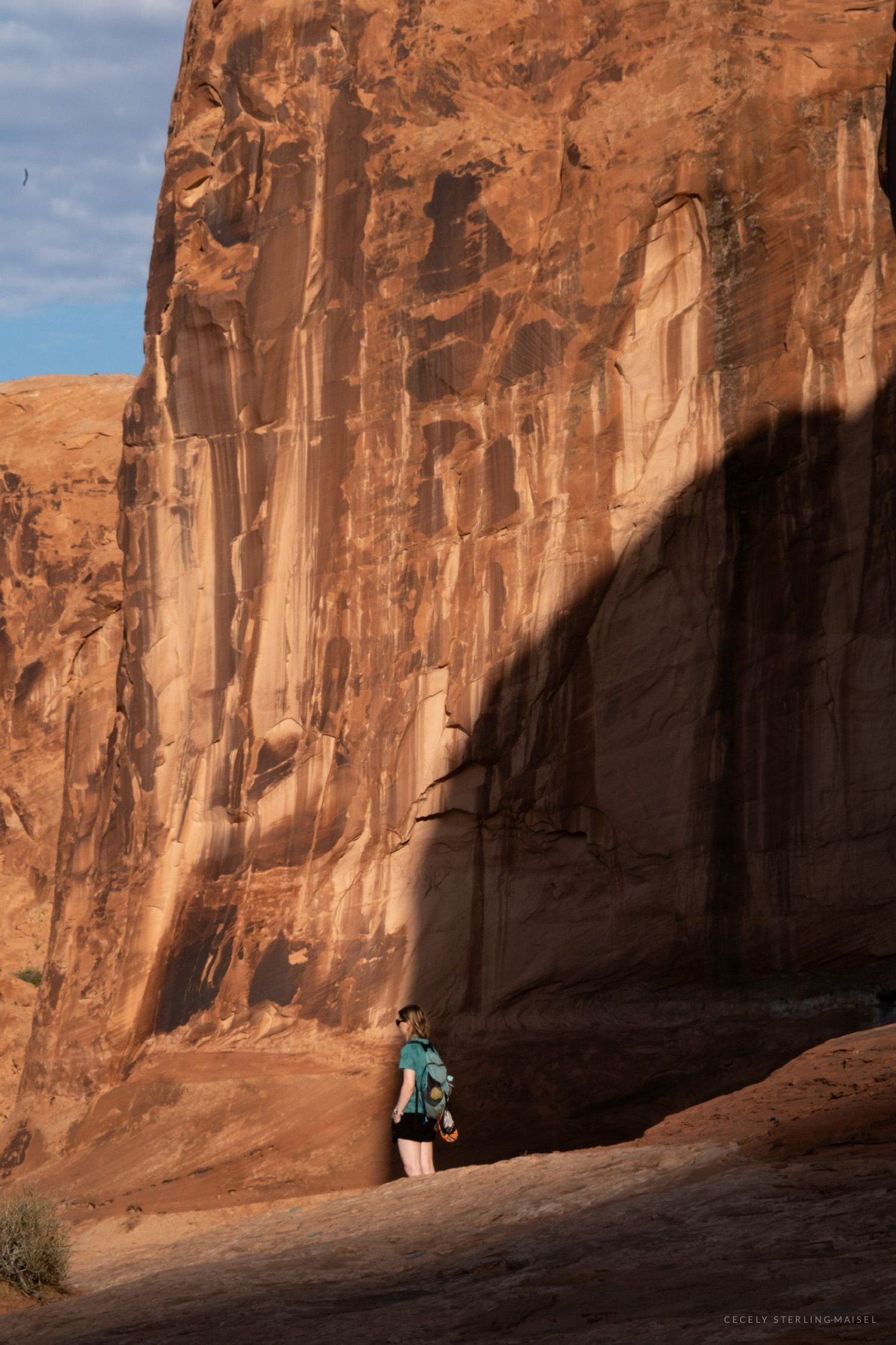A small figure in a teal shirt standing at the foot of an immense sunlit sandstone wall, a curved shadow falling across it.