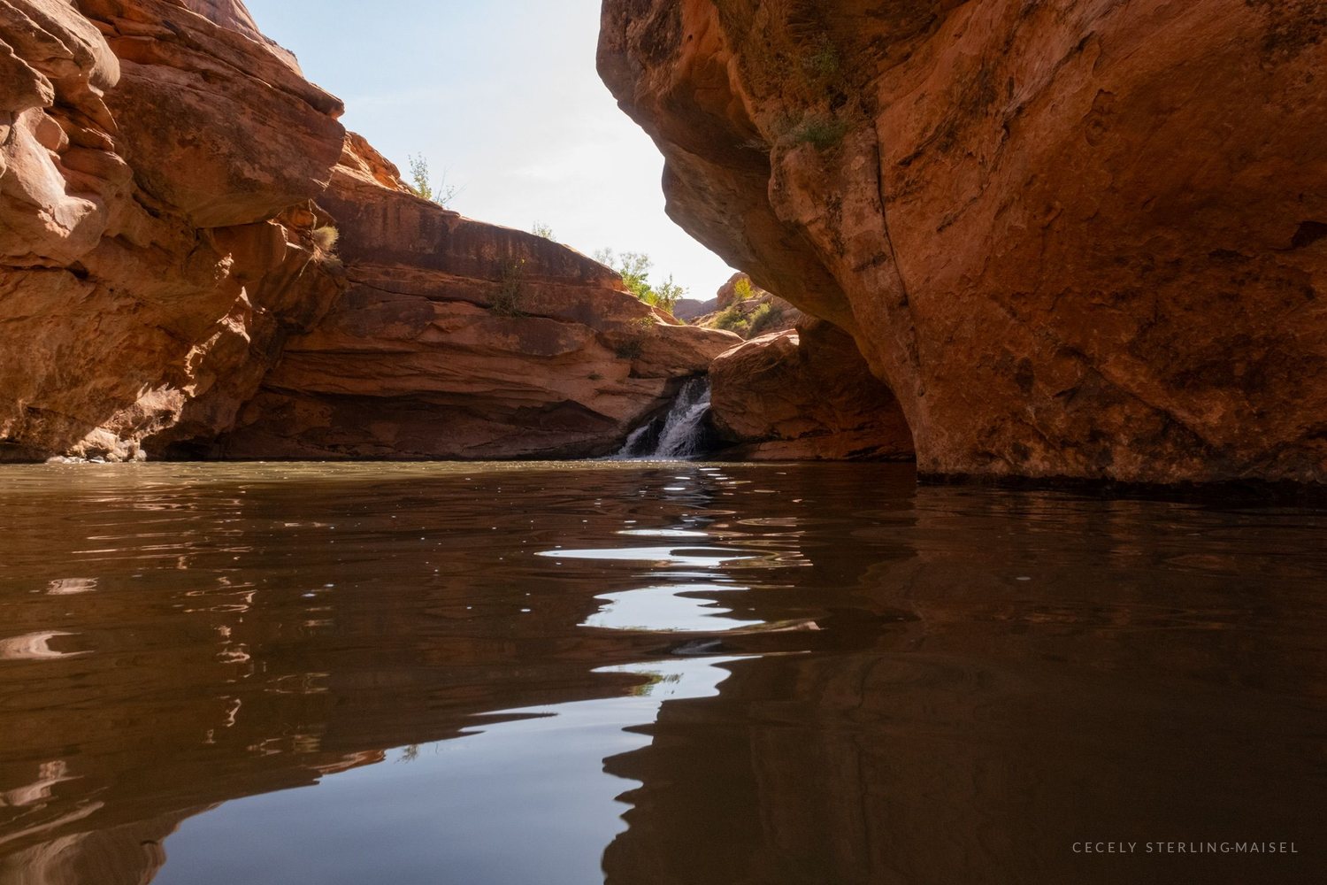 A shallow waterfall spilling into a still canyon pool, the sky reflected in a band down the middle of the water.