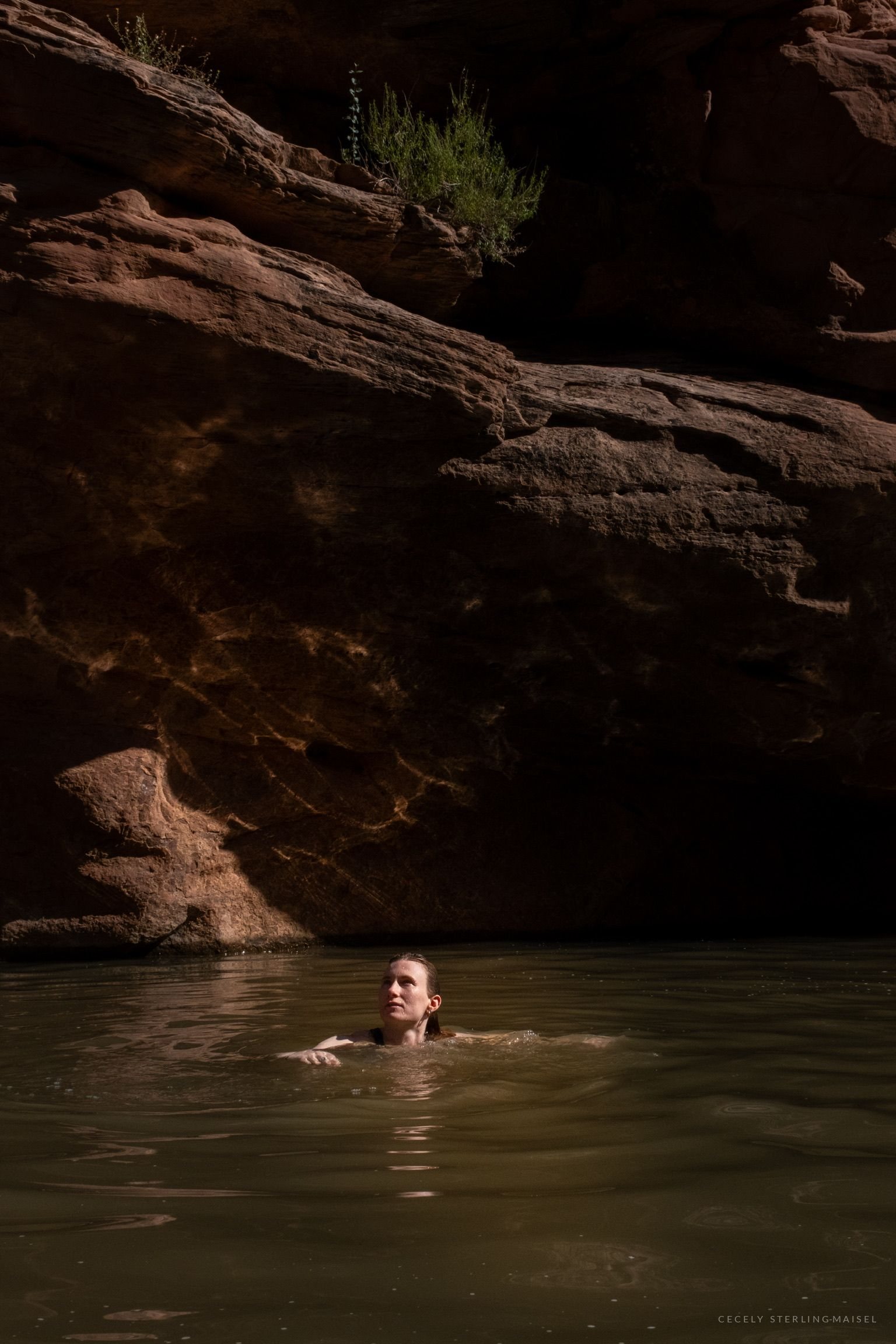 A swimmer's head above dark green water beneath a huge overhanging shelf of banded sandstone.