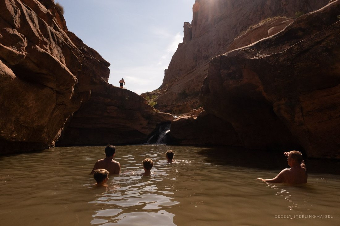 Four swimmers in a green canyon pool below a waterfall, a lone figure standing on the rim far above.