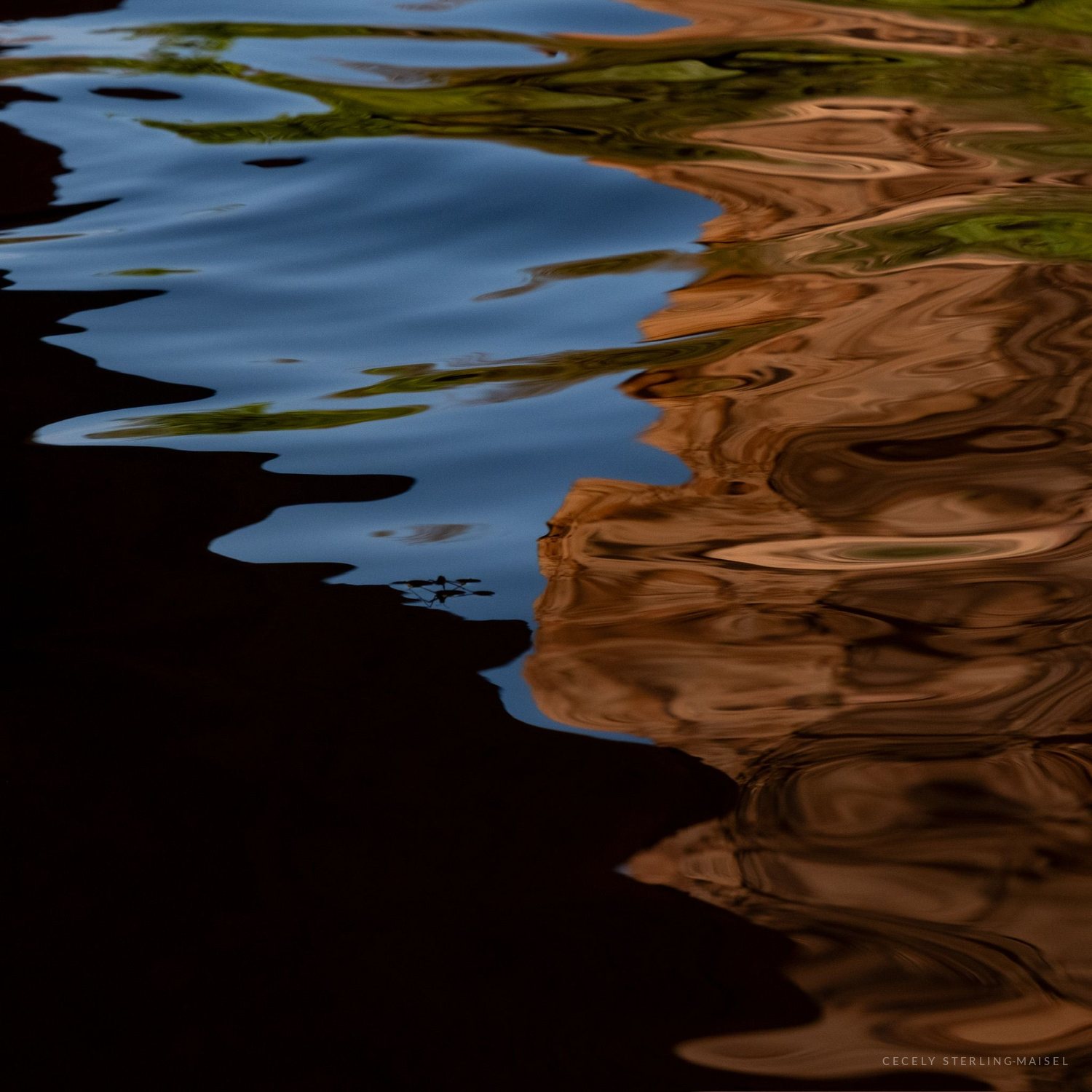 Blue sky and green trees reflected in rippling water, breaking into bands of ochre and black.