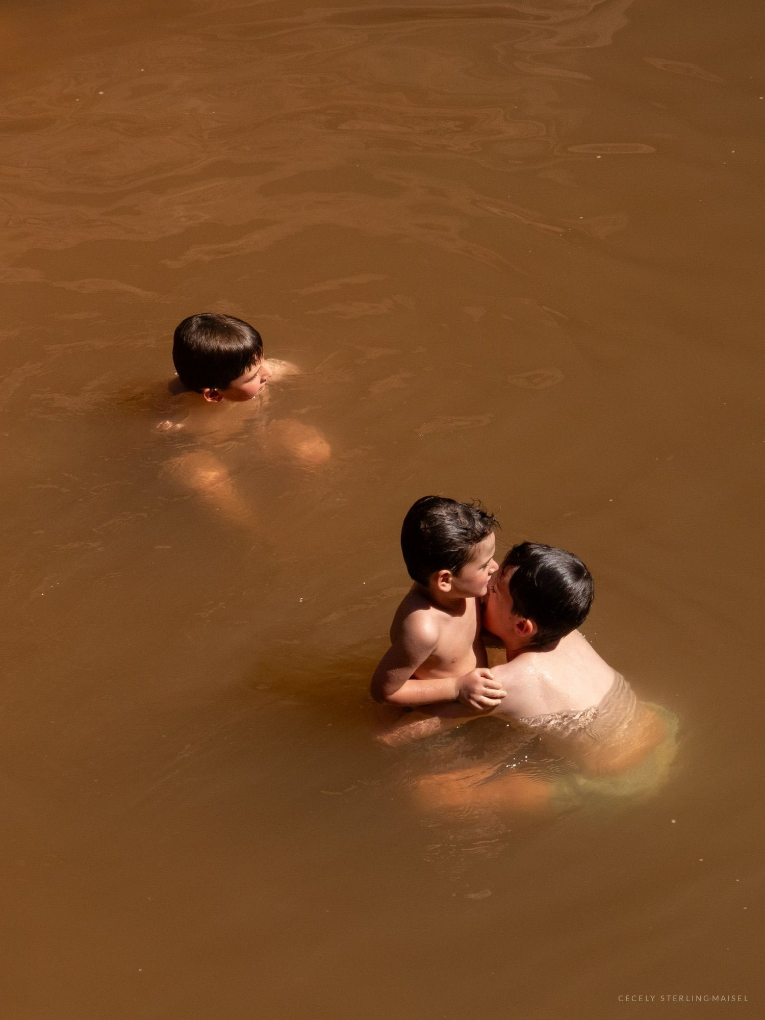 Three young people standing chest-deep in ochre water seen from above, two of them leaning together.
