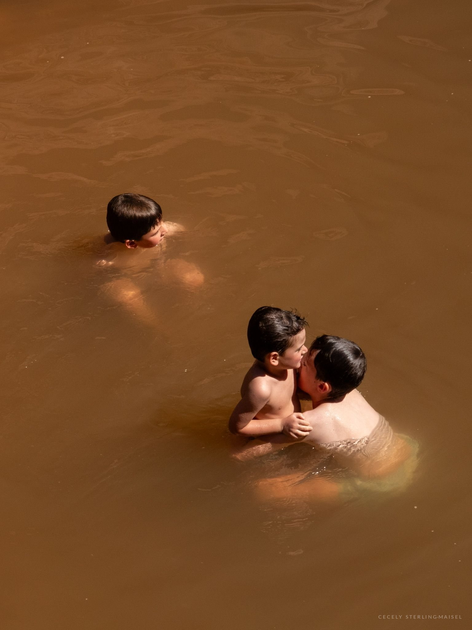 Three young people standing chest-deep in ochre water seen from above, two of them leaning together.