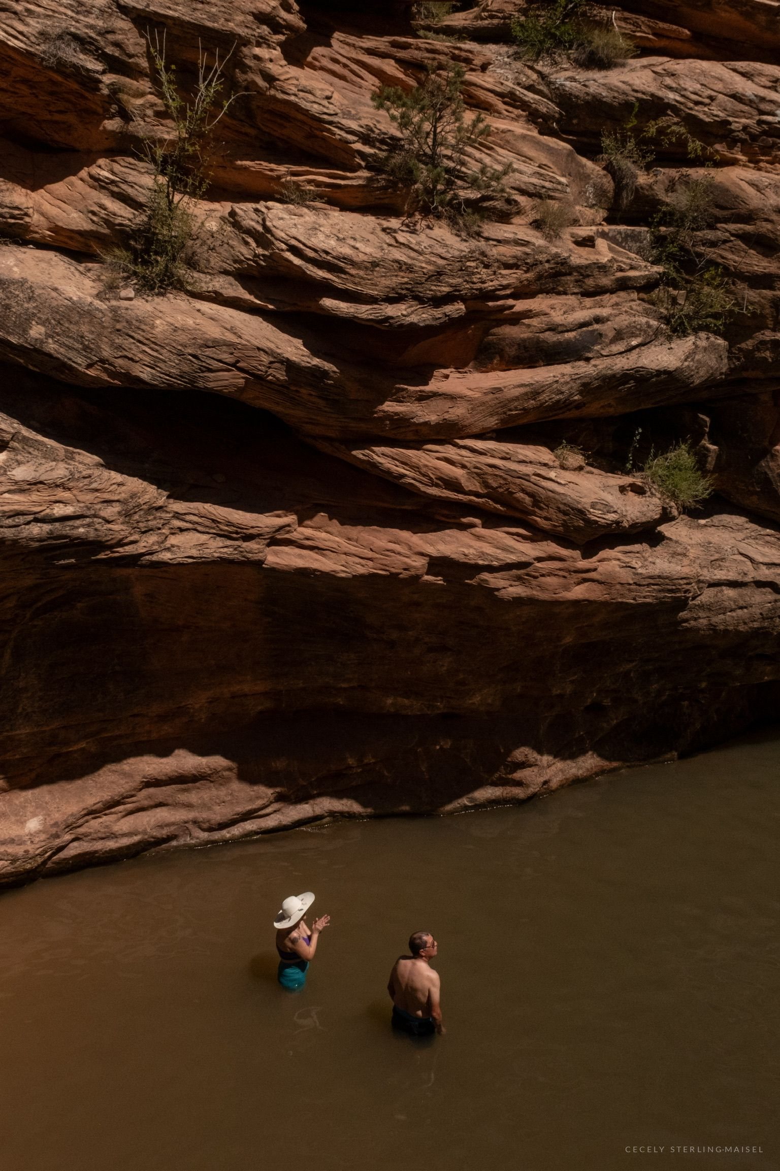 Two small figures wading in a muddy pool at the foot of a broad tier of sandstone ledges.