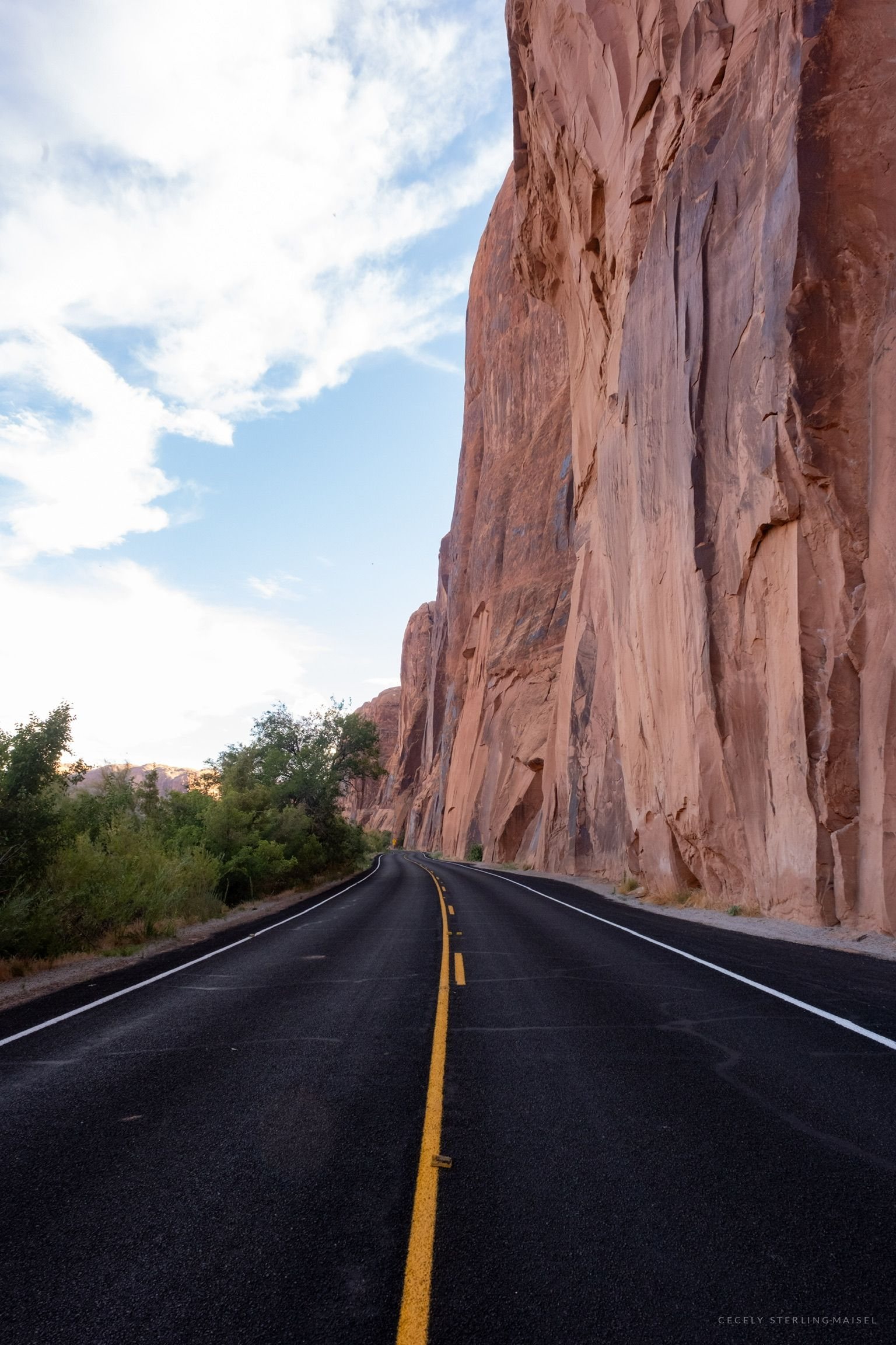 A road running along the foot of a sheer sandstone cliff, the yellow centreline leading into the distance.