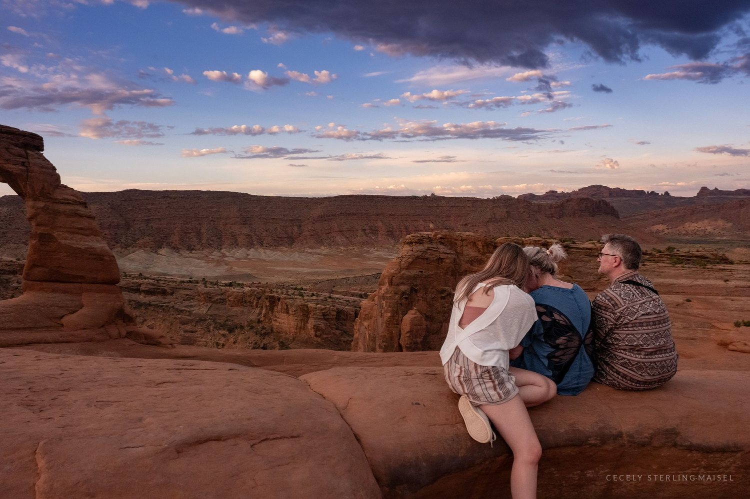 Three people sitting together on slickrock watching Delicate Arch under a pink and blue evening sky.
