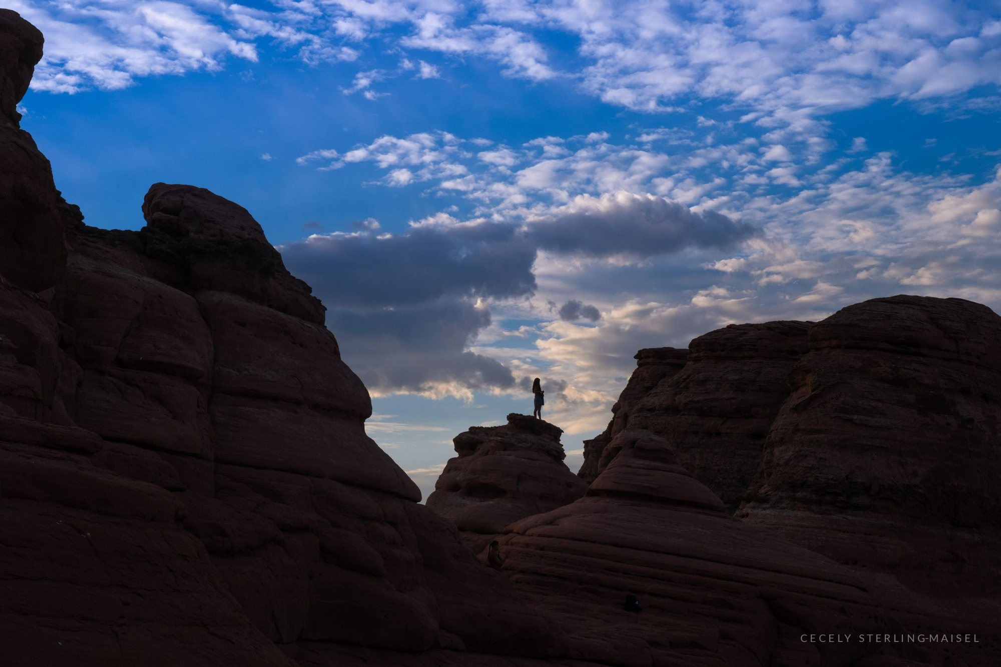 A lone figure standing on a sandstone fin, silhouetted against towering cloud.