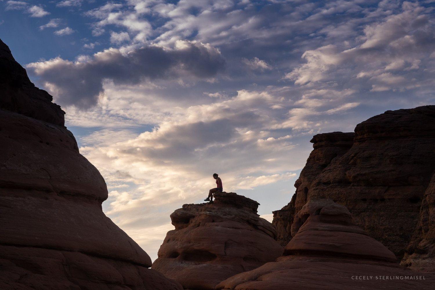 A person sitting on top of a rounded sandstone dome as the sun goes down behind broken cloud.