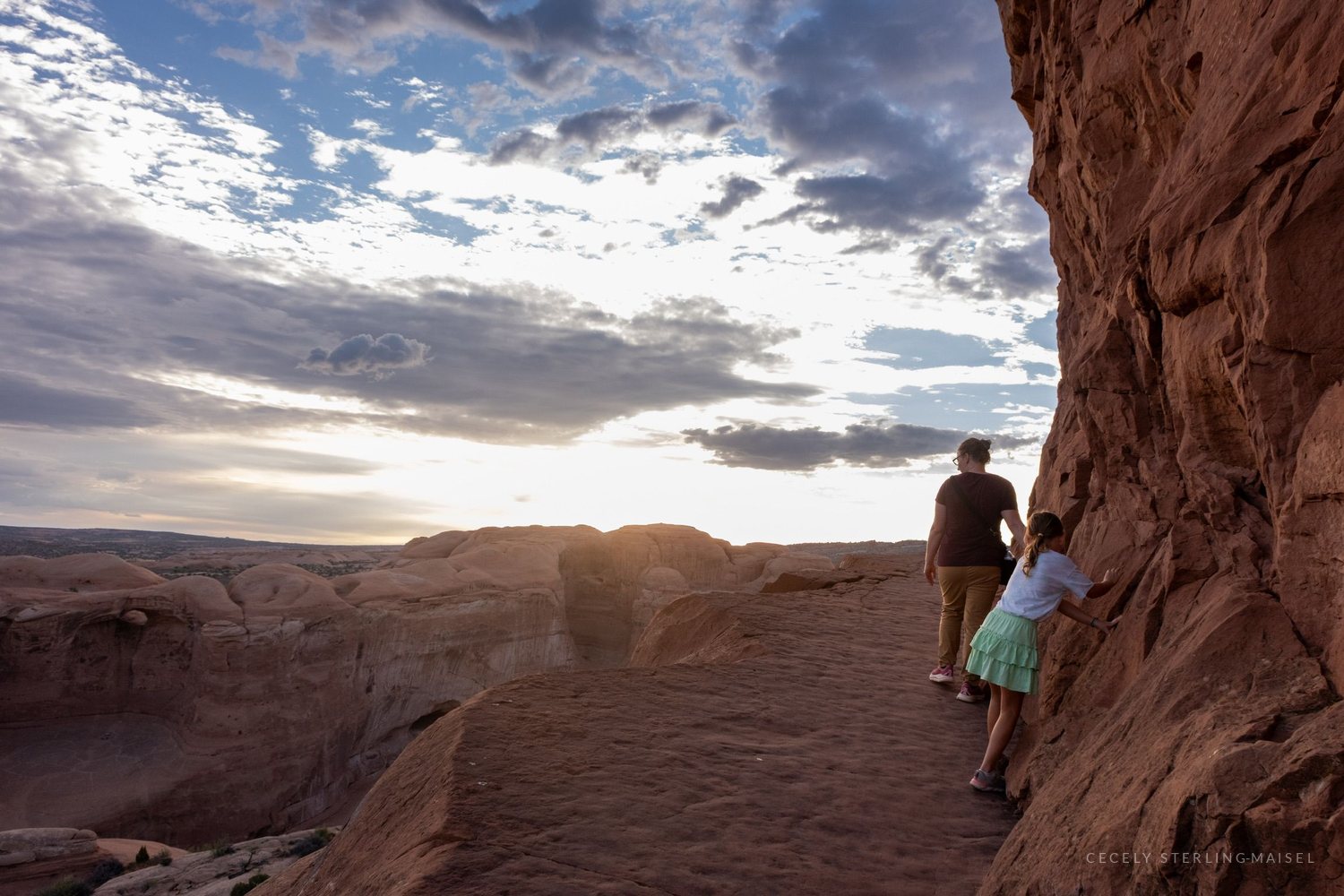 A man and a child at the edge of a slickrock bowl, the low sun flaring around the rock beside them.