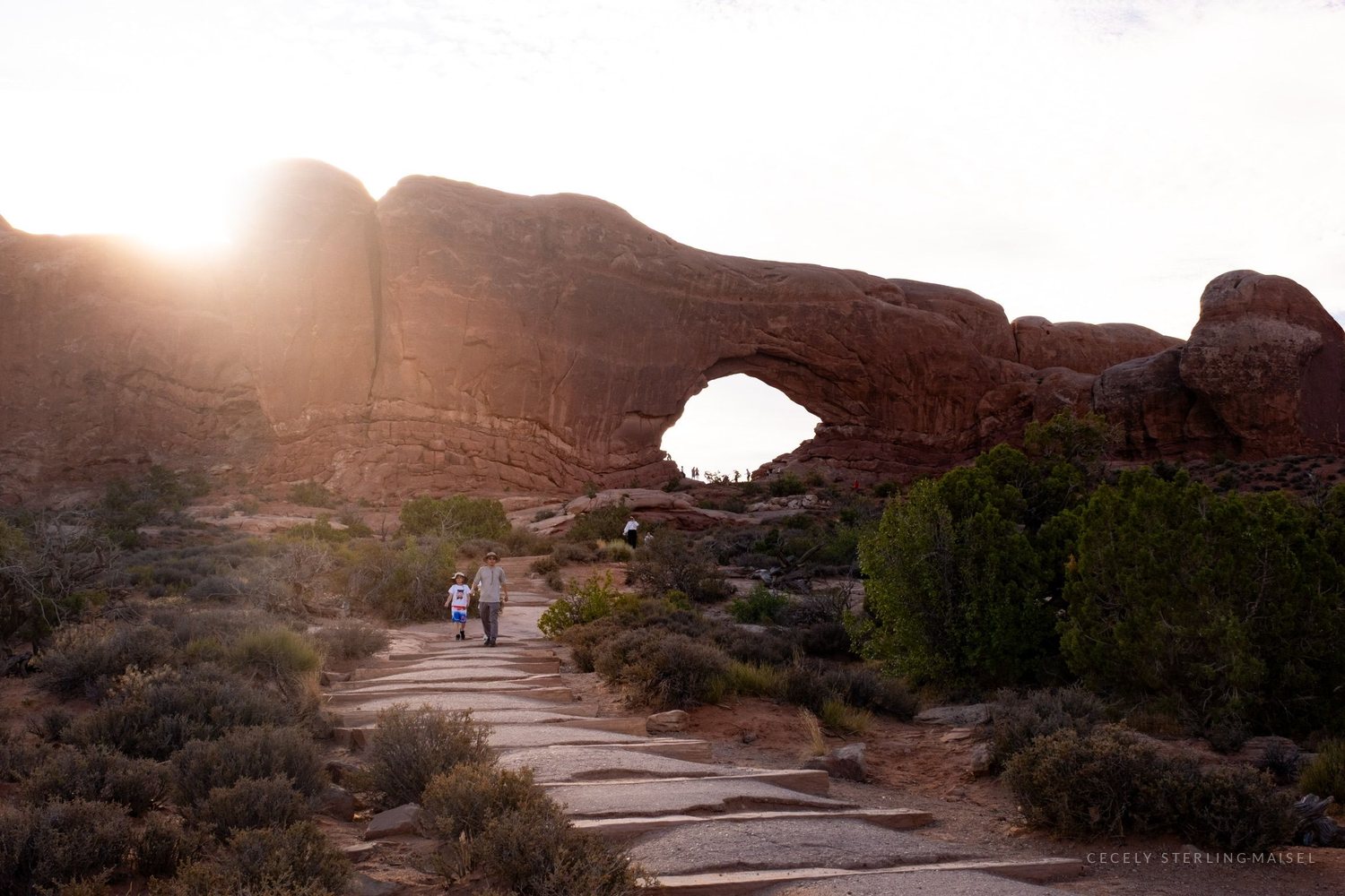 A stone path leading toward North Window arch with the sun breaking over the rim and two figures on the trail.