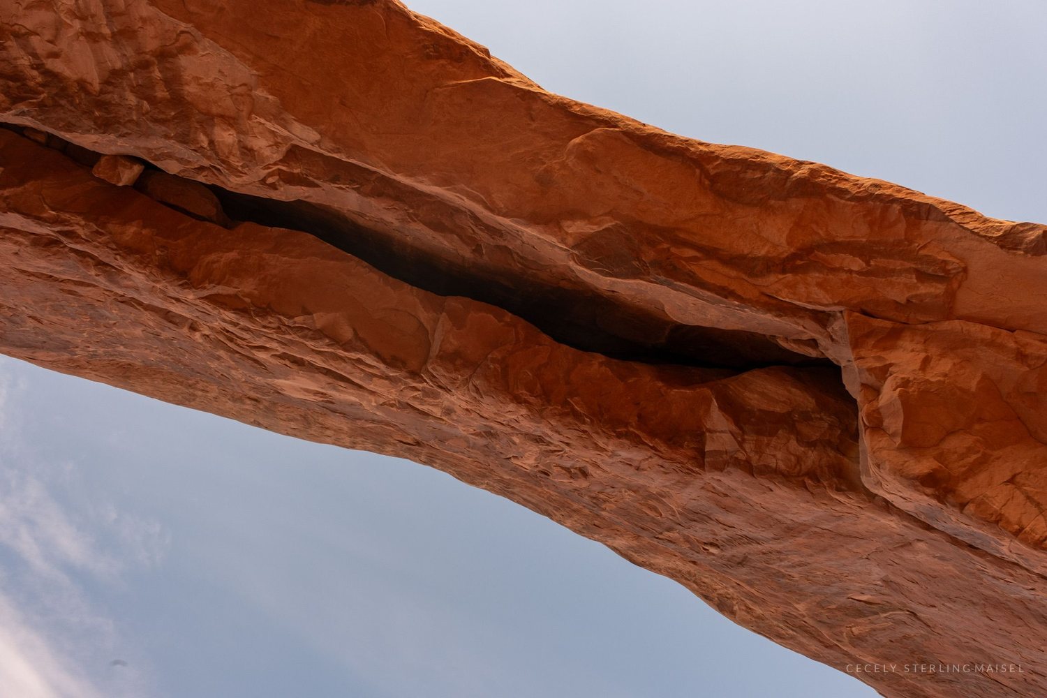The underside of a long sandstone arch lit orange, cutting diagonally across a pale sky.