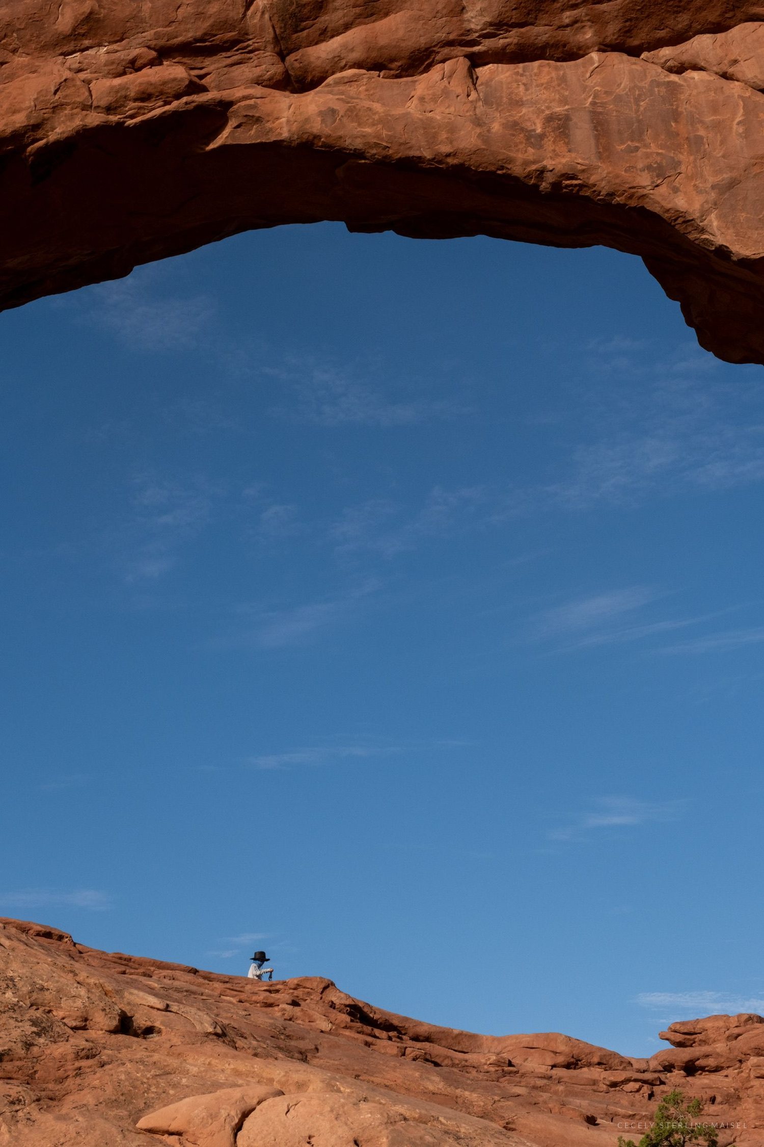 A wide arch seen from beneath framing blue sky, with a tiny figure standing on the rock below it.