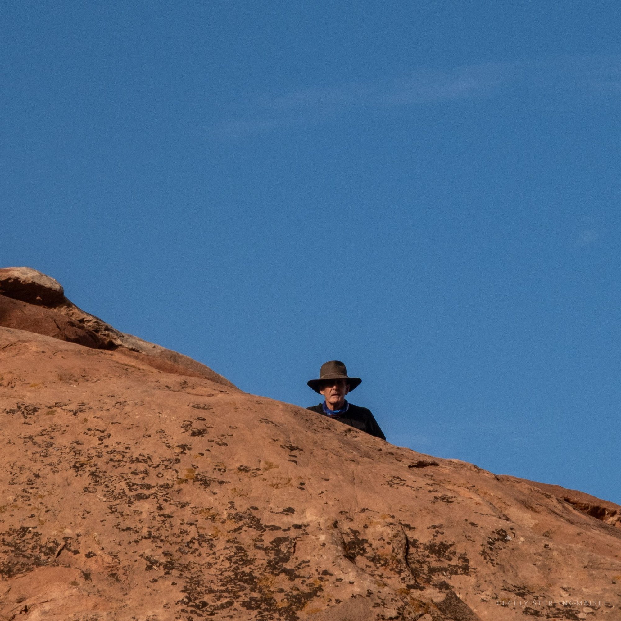 A man in a wide-brimmed hat just visible over the crest of a sandstone slope against deep blue sky.