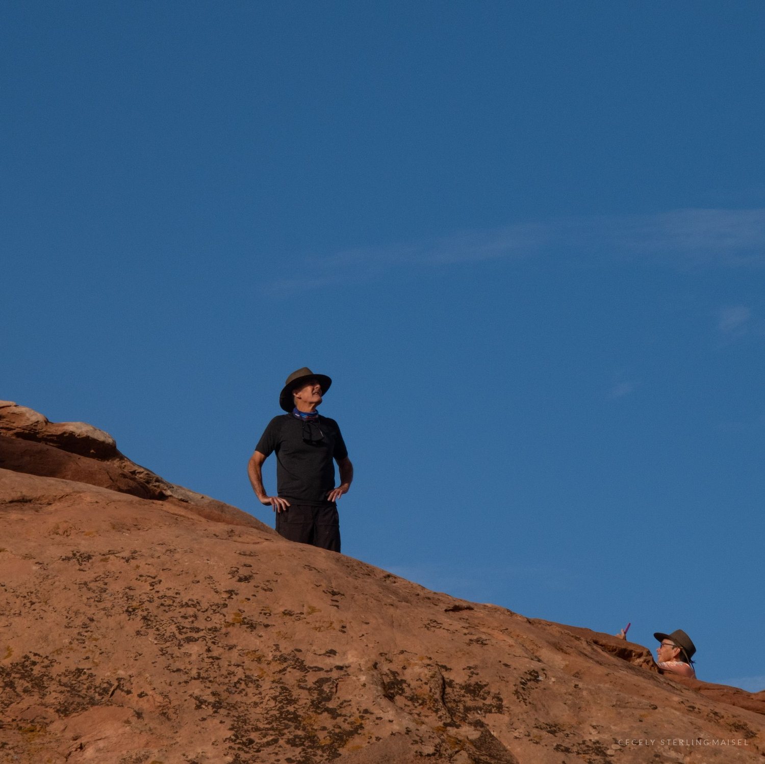A man in a hat standing on a bare sandstone slope, a second figure's head appearing over the crest below him.