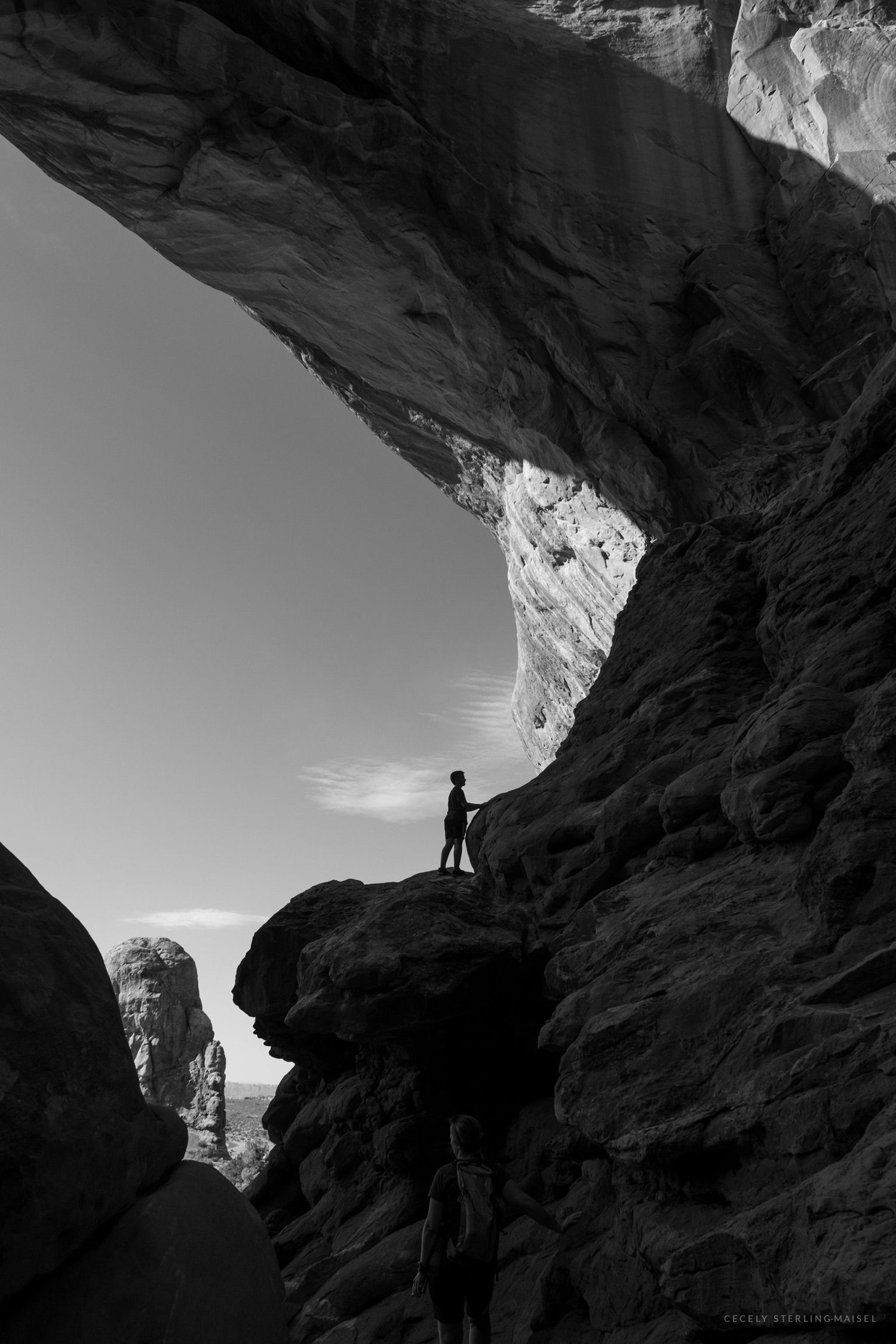 Black and white photograph of a figure standing on a rock shelf under a dark overhang, bright rock fins beyond.