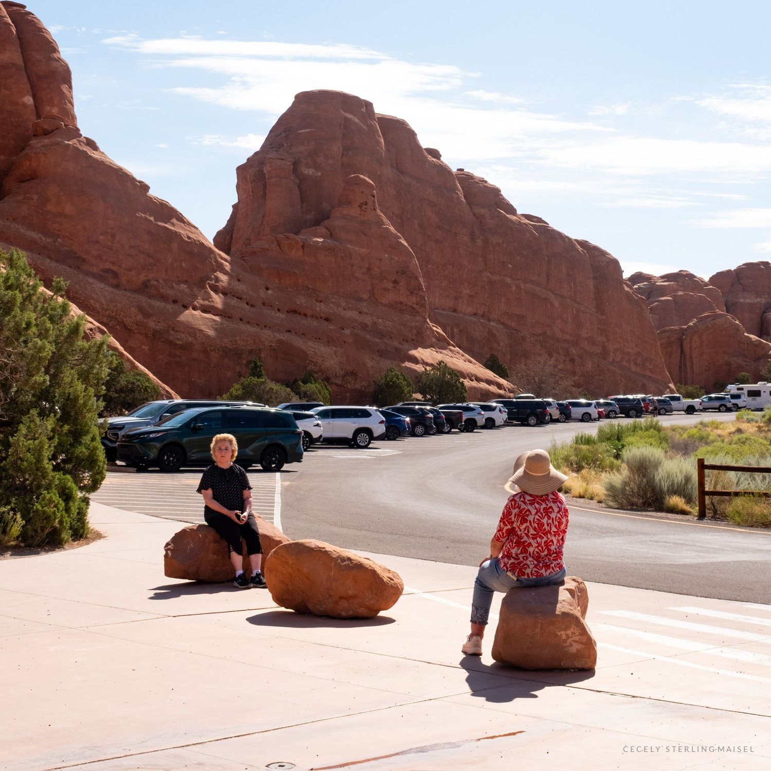Two people sitting on boulders beside a car park at the edge of the red rock at midday.
