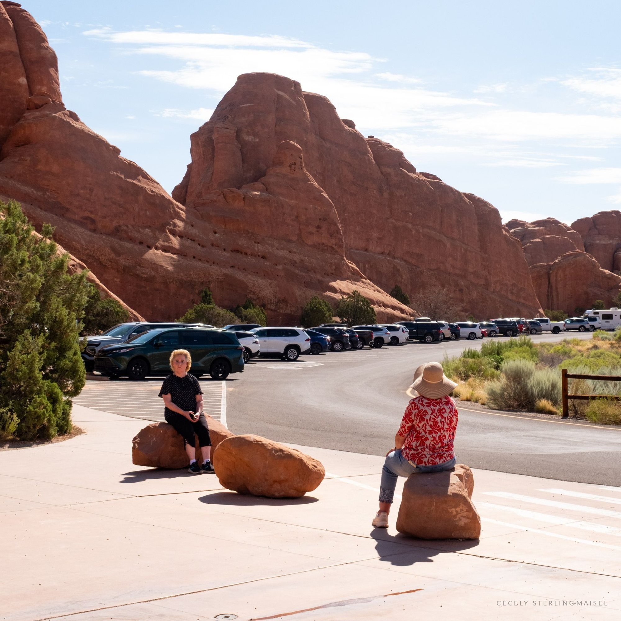 Two people sitting on boulders beside a car park at the edge of the red rock at midday.
