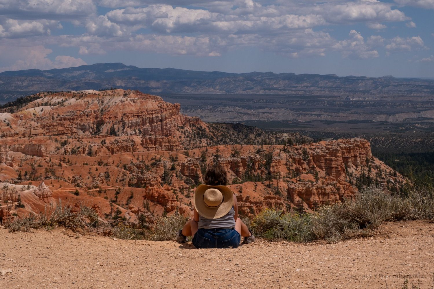 A person in a straw hat sitting alone on the rim looking out across the canyon to distant plateaus.