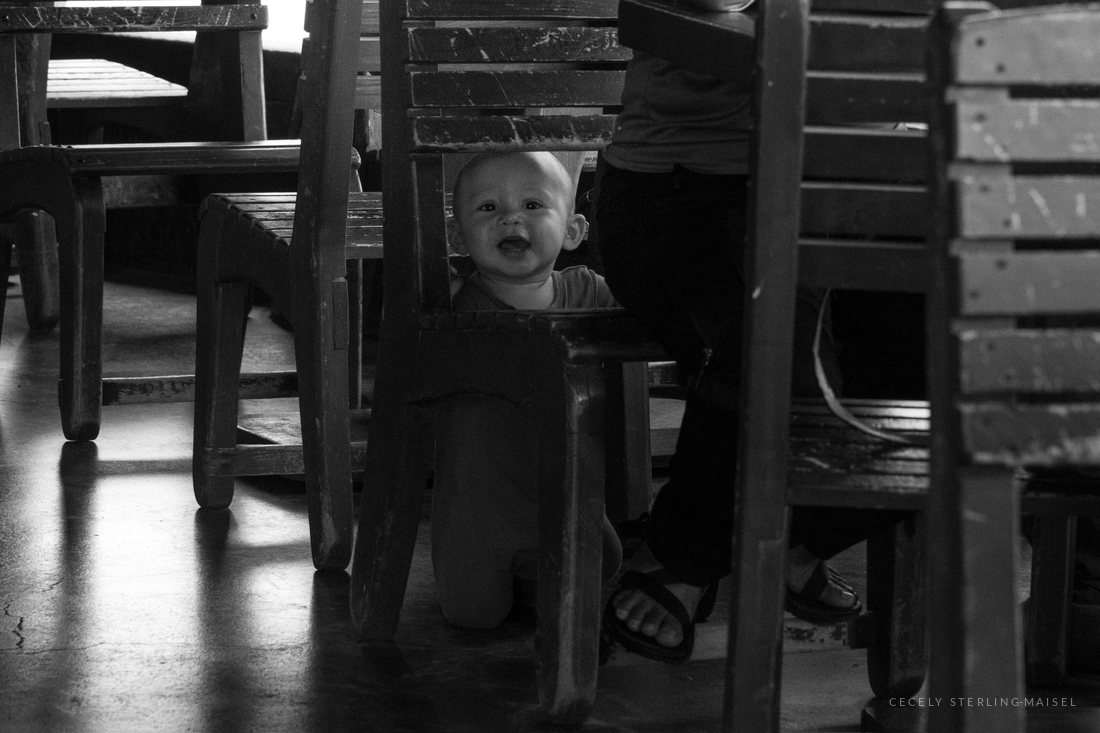 A baby sitting on the floor between the legs of wooden chairs and tables, looking up and laughing.