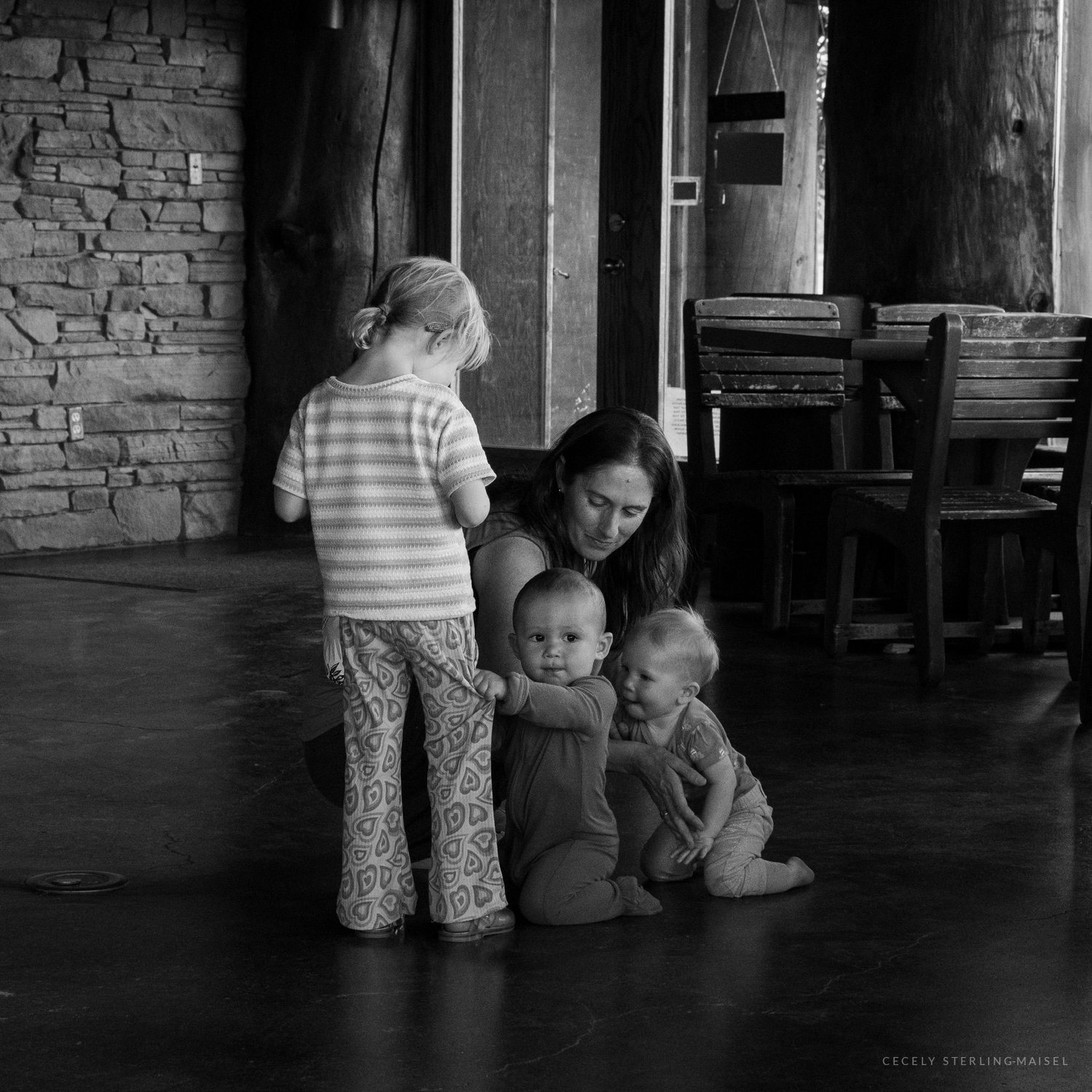 Black and white photograph of a woman crouched on a lodge floor with three small children gathered around her.