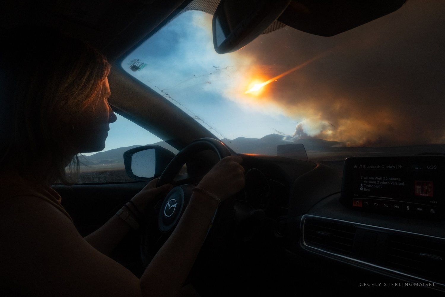 A woman driving with one hand on the wheel, a wildfire burning across the valley beyond the windscreen.