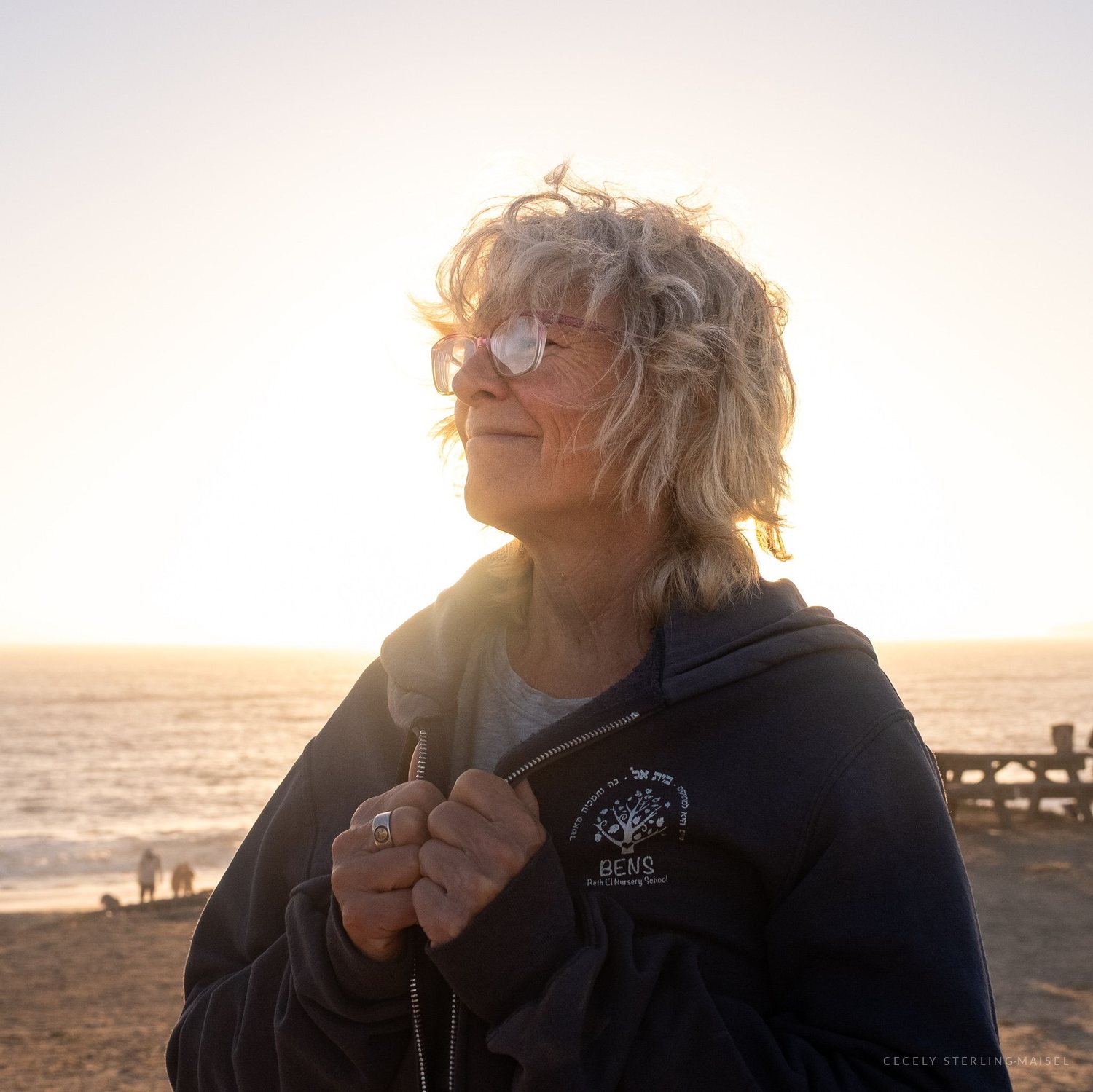 An older woman with white hair standing on a beach at sunset, eyes closed, the light burning out behind her.