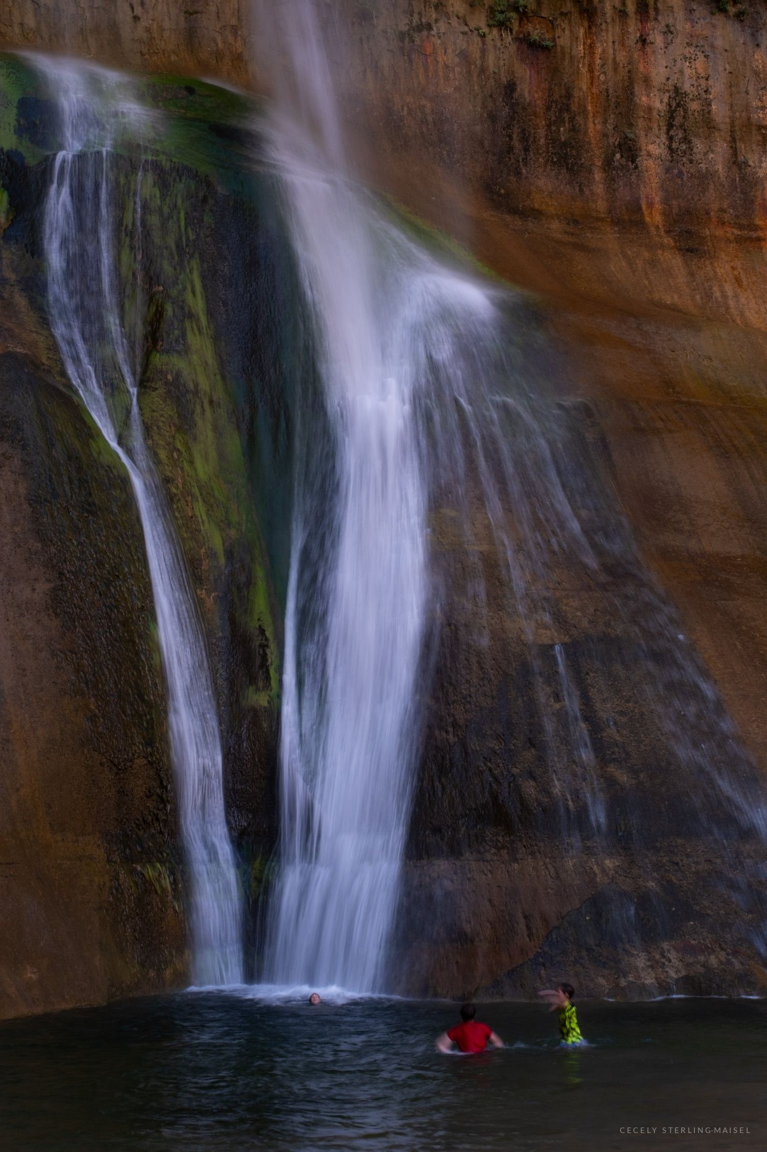 Two swimmers in the plunge pool beneath a tall desert waterfall falling down a streaked sandstone wall.
