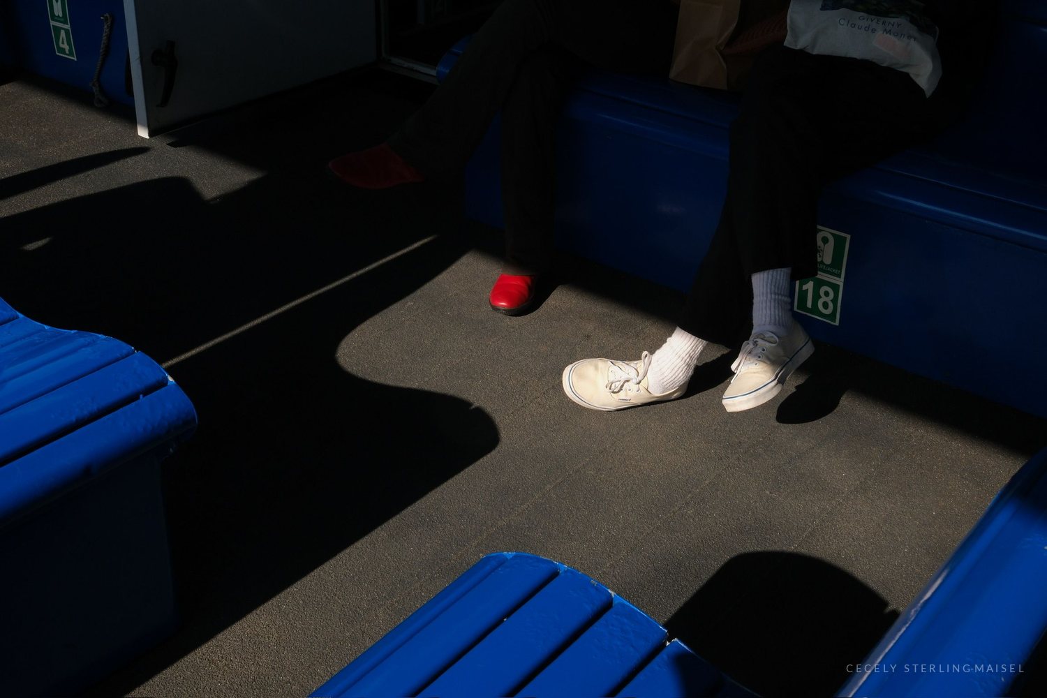 Sunlight falls across the deck of a ferry, catching a passenger's white shoes and socks between rows of blue seats.