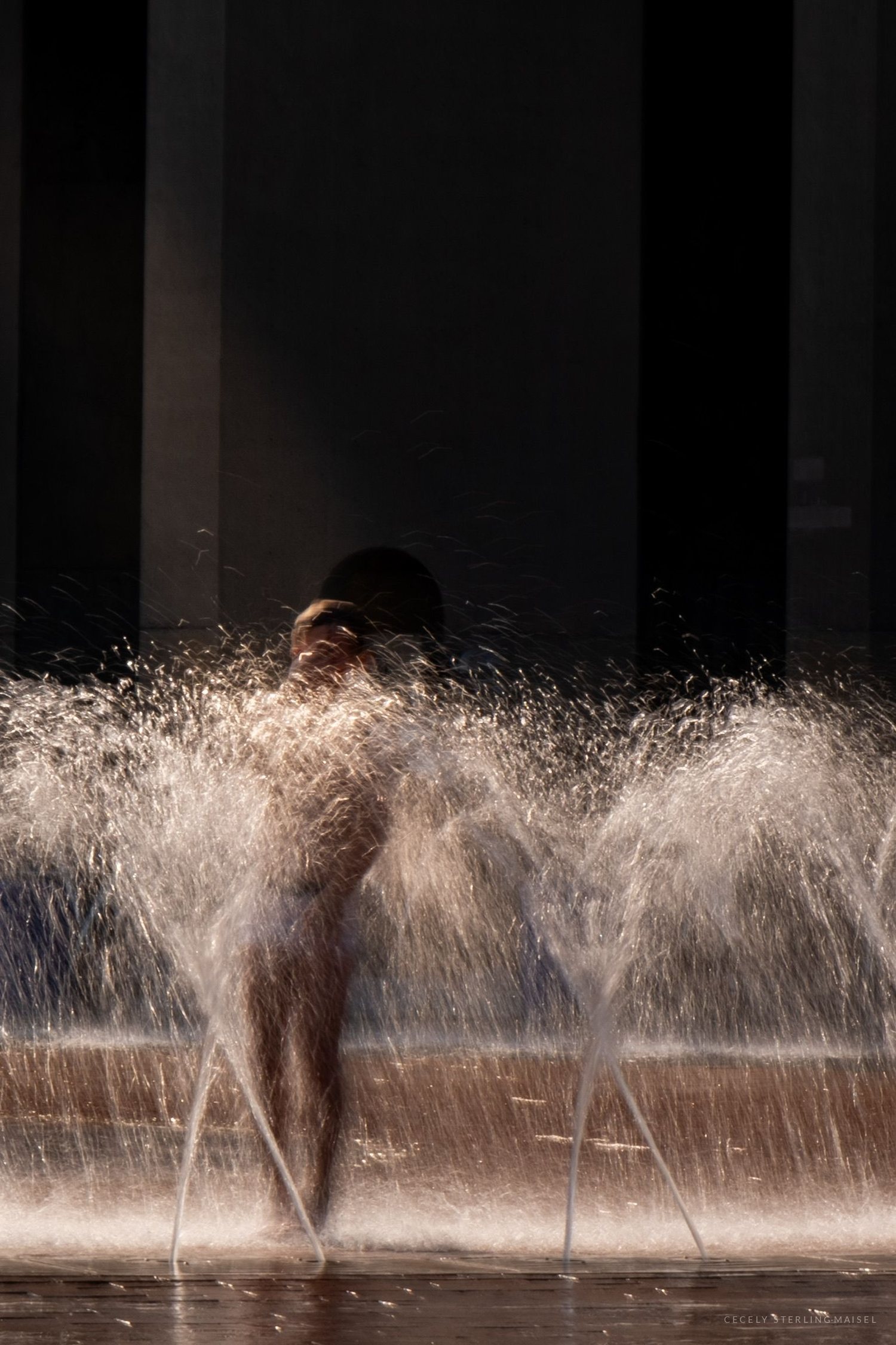 A figure stands in the arcing jets of a city fountain, backlit against a dark building.