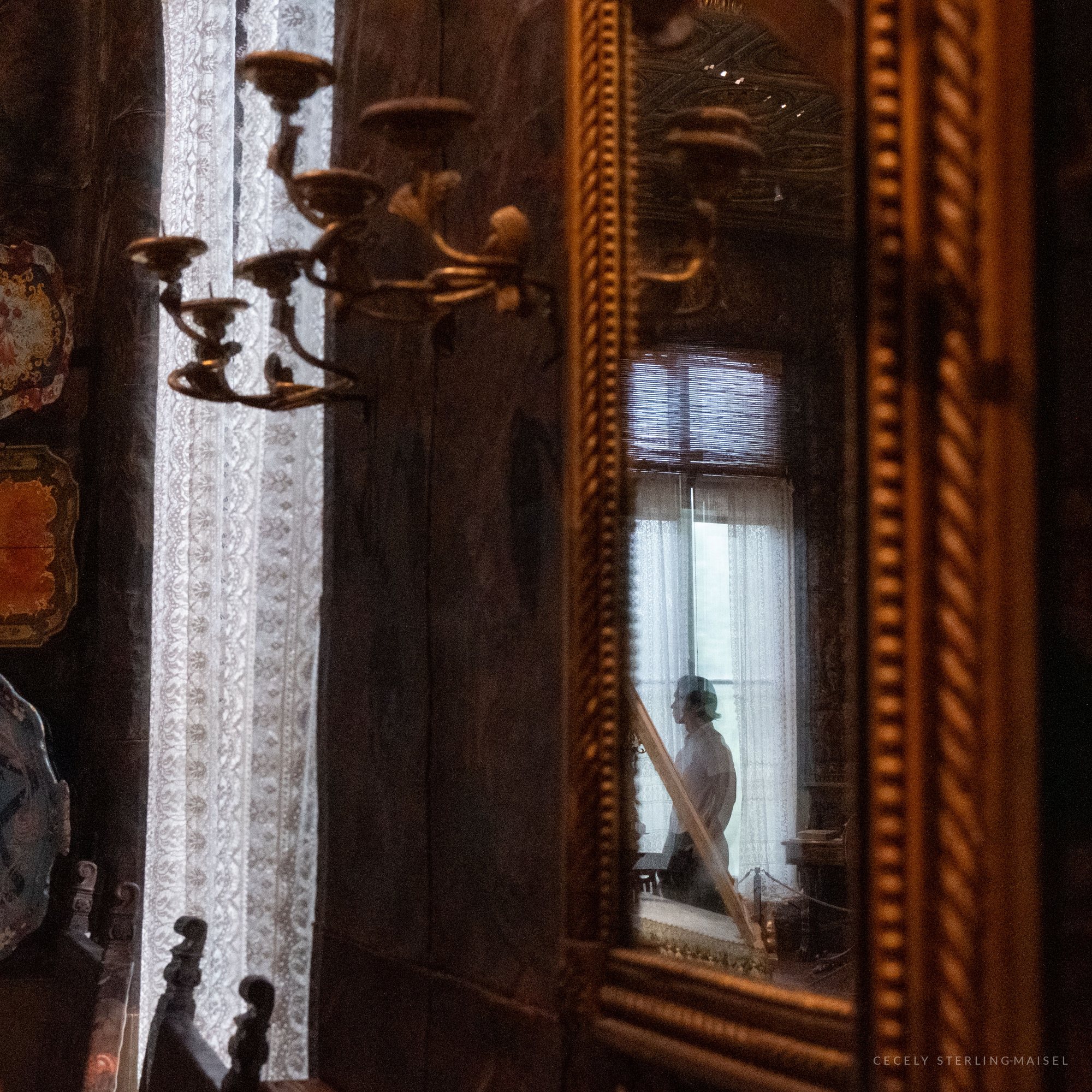 A gilt mirror in a dim, antique-filled room reflects a person standing at a lace-curtained window, with a brass candelabra in the foreground.