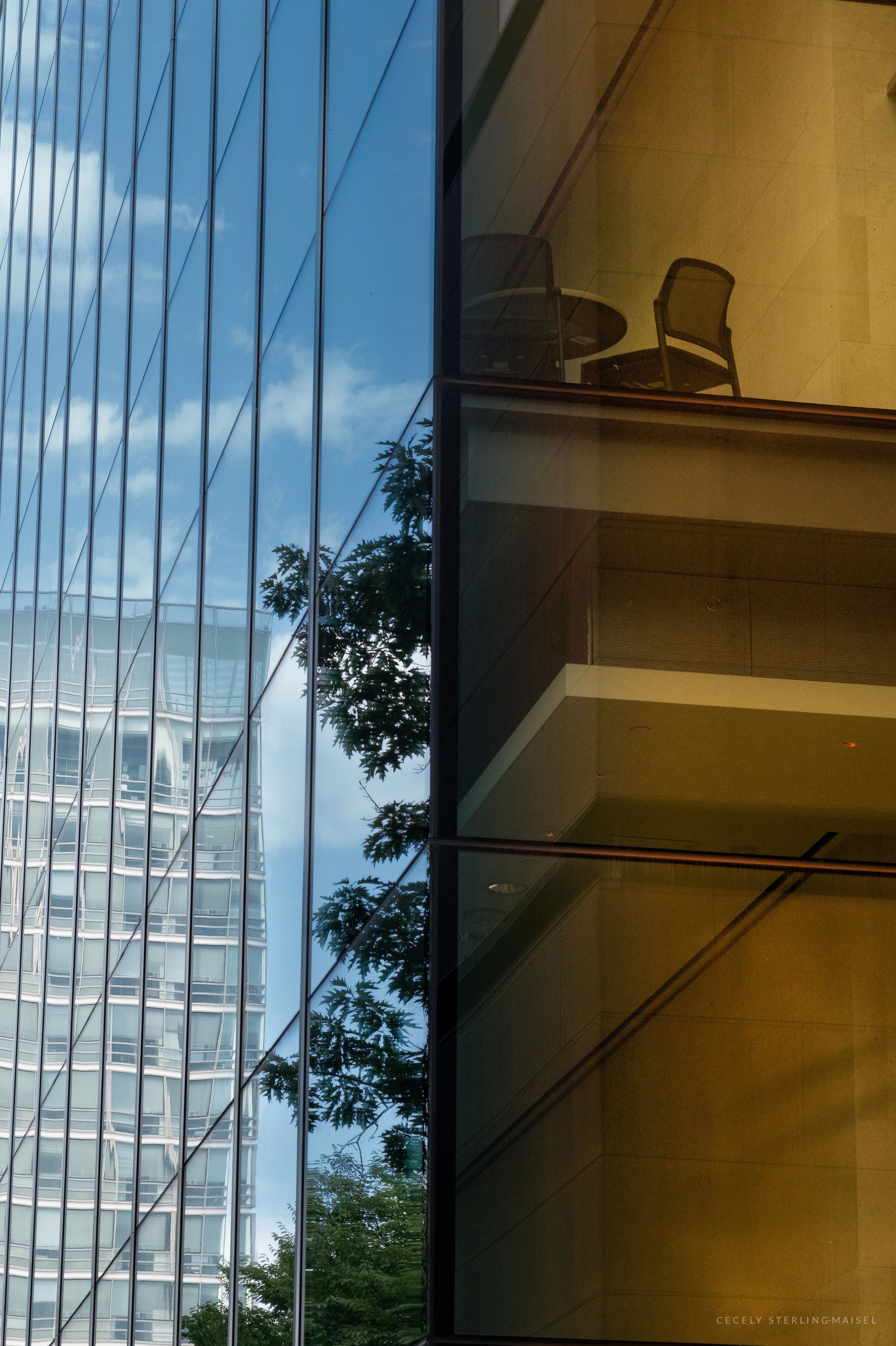 A glass office facade at an angle, one side mirroring blue sky, clouds, a tree and a neighboring tower, the other revealing a lit interior with a small table and chairs.