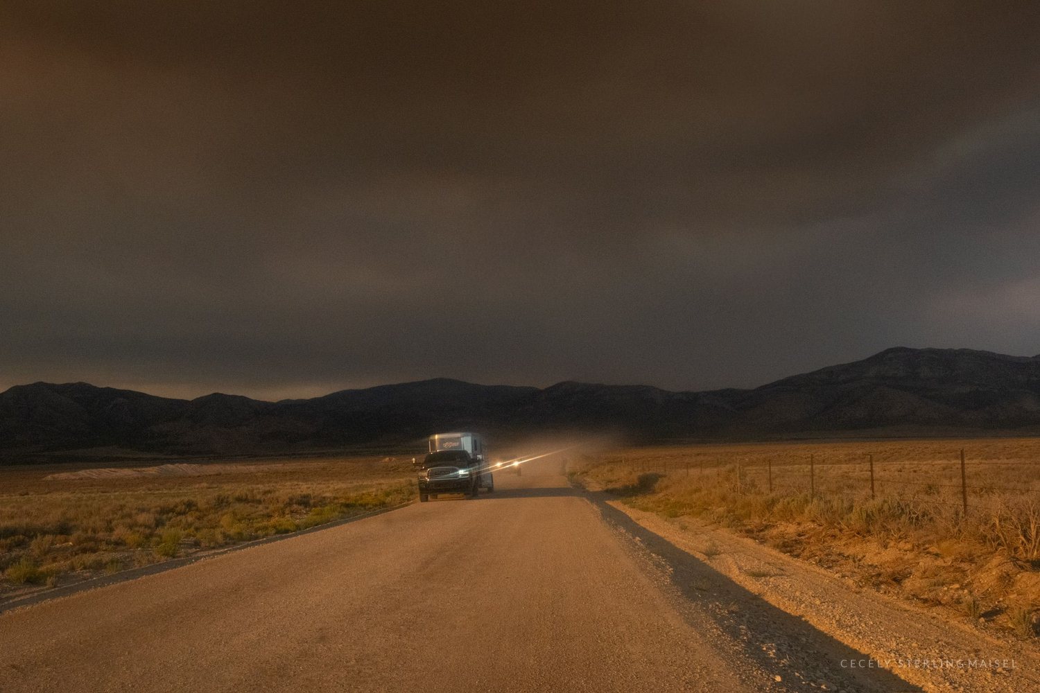 A truck's headlights on an empty desert road at dusk beneath a low ceiling of wildfire smoke.