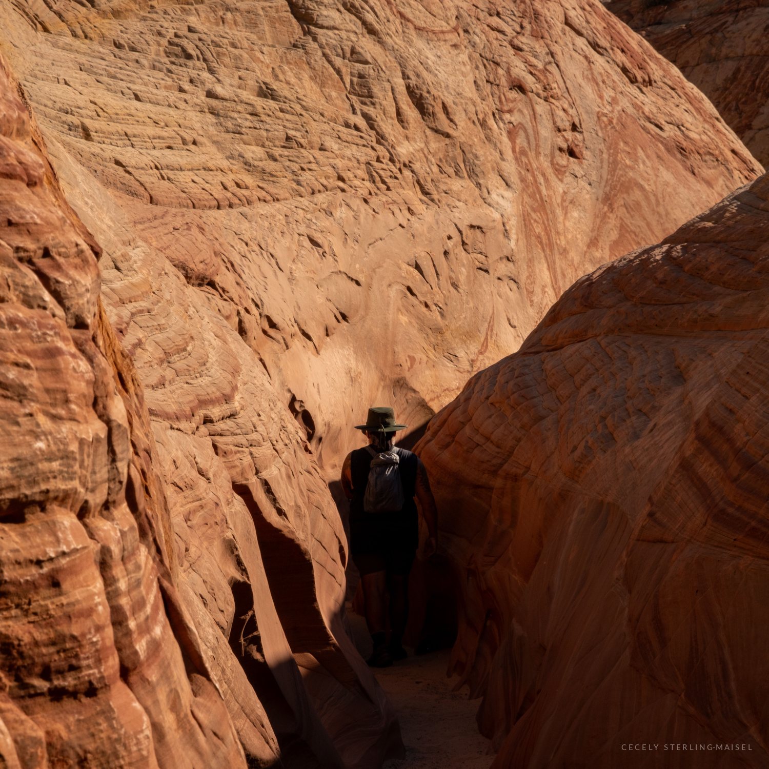 A hiker in a green hat walks through a narrow sandstone slot canyon, silhouetted against swirling orange and cream rock walls