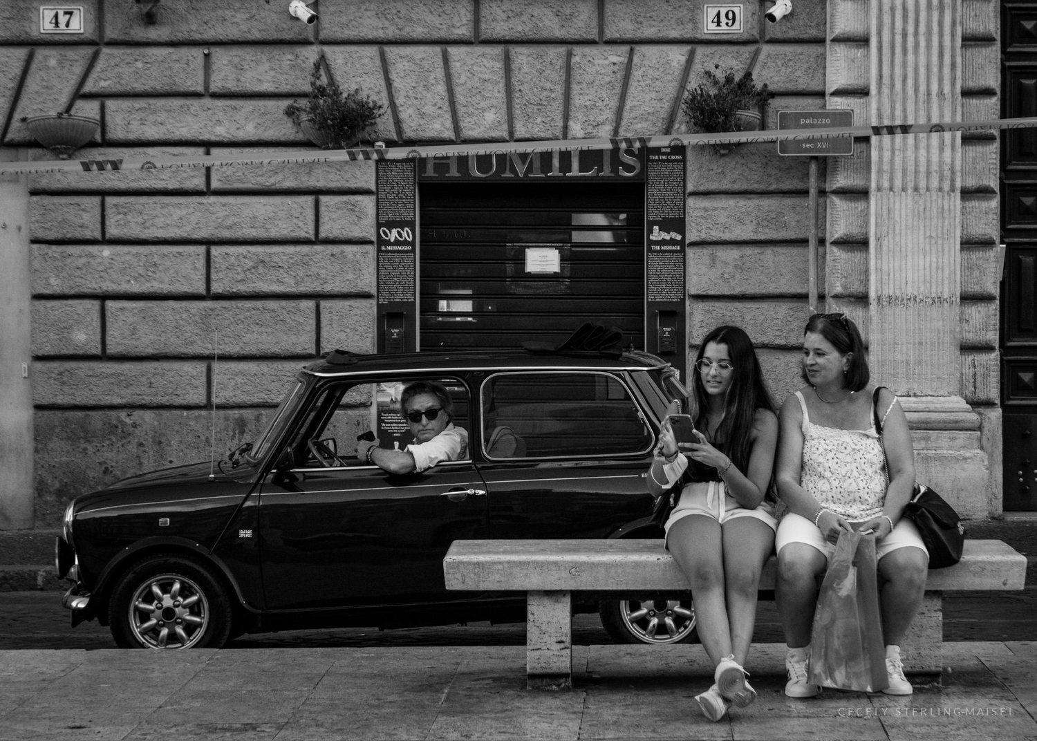 Black and white photograph of two young women seated on a bench beside a classic Mini, a man watching from the driver's seat.