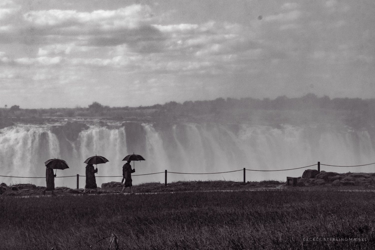 Black and white photograph of three figures with umbrellas walking a fence line in front of a vast waterfall.