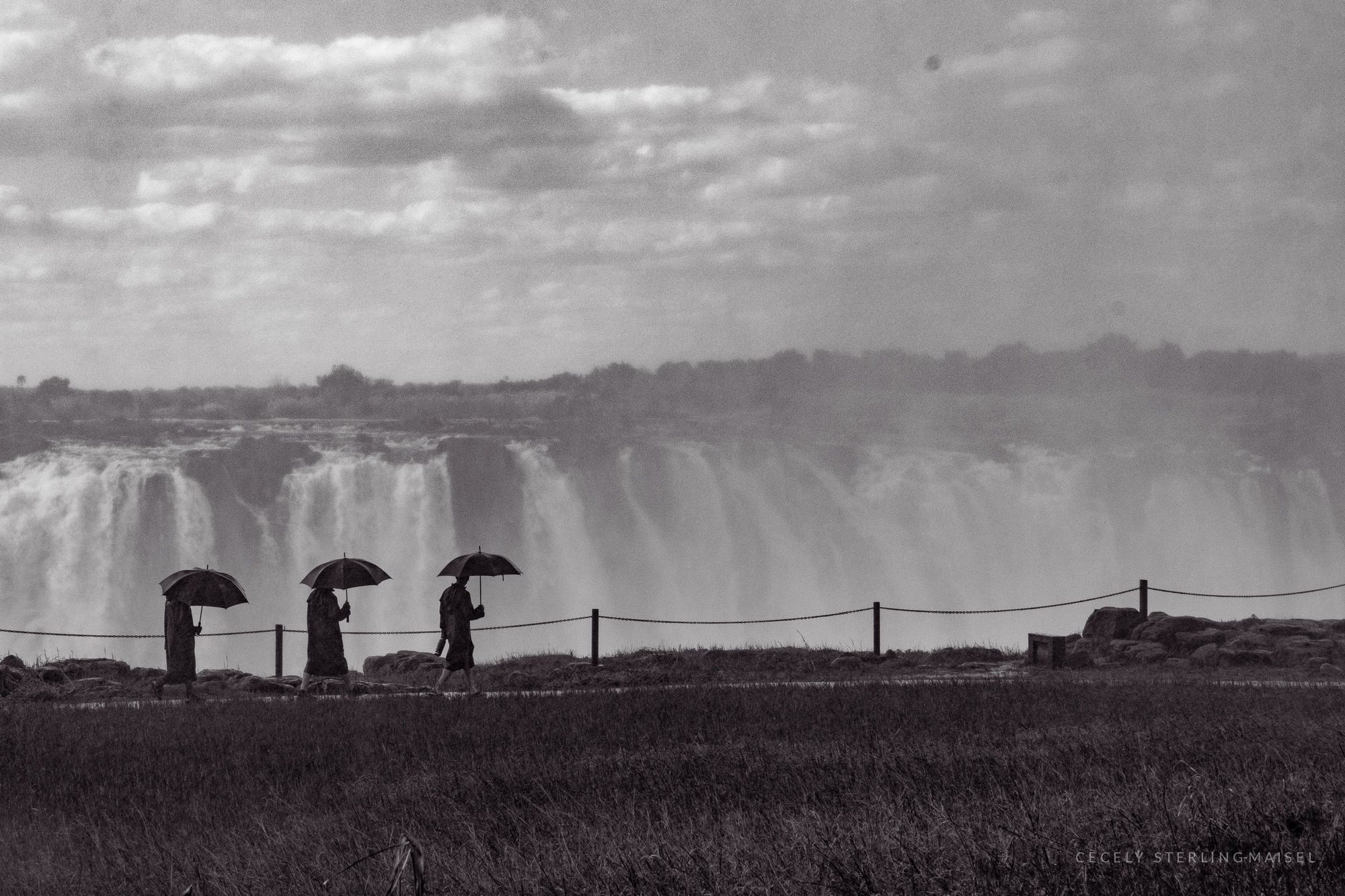 Black and white photograph of three figures with umbrellas walking a fence line in front of a vast waterfall.